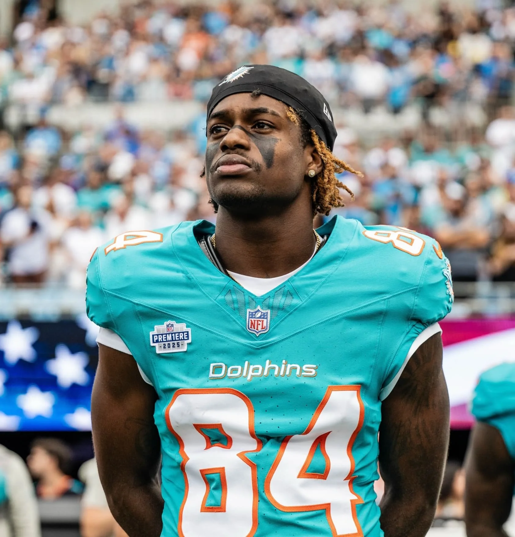 A football player in a teal Miami Dolphins jersey with the number 84, wearing a black headband, standing on the field at a stadium with a blurred crowd in the background.