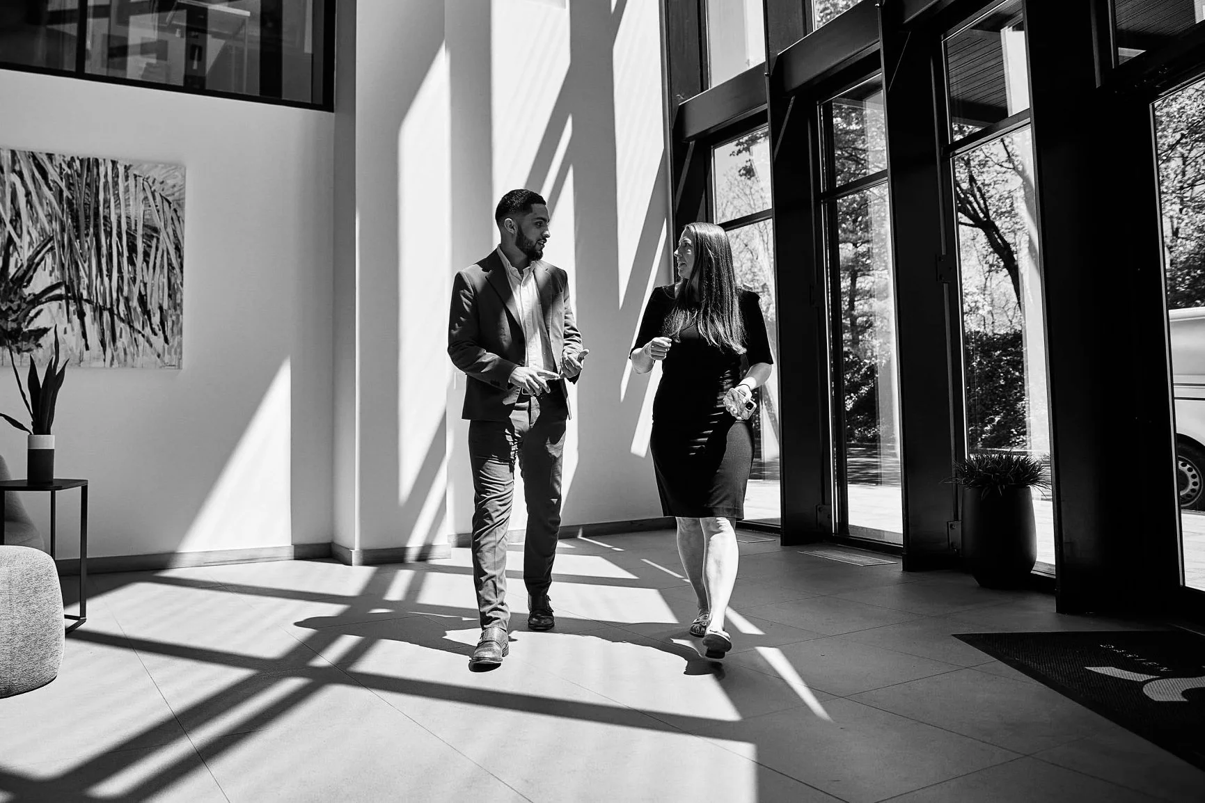 Two people, a man in a suit and a woman in a dress, are walking and talking inside a modern building with large windows. Sunlight casts shadows across the floor.