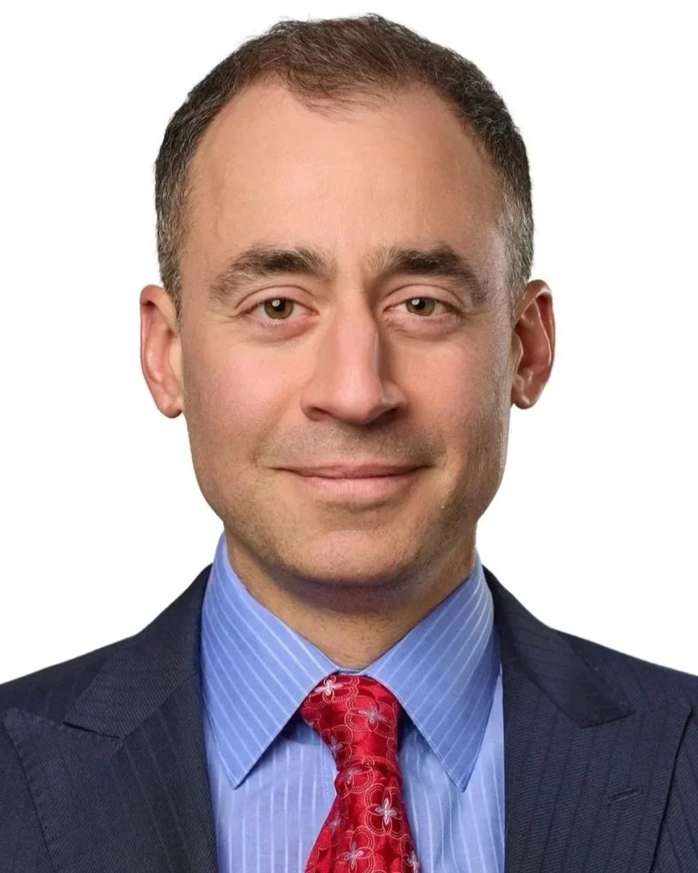 A professional headshot of a man with short brown hair, wearing a dark suit, light blue dress shirt, and red patterned tie, against a white background.