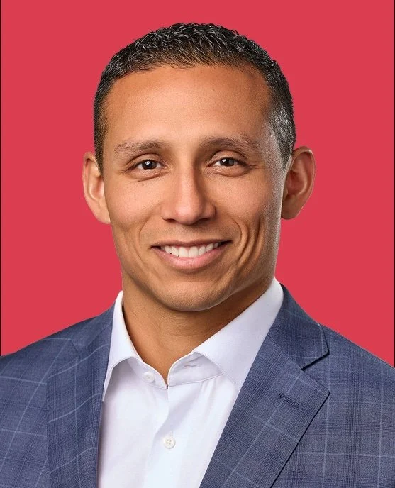 Professional man wearing a blue suit and white shirt, smiling against a pink background.