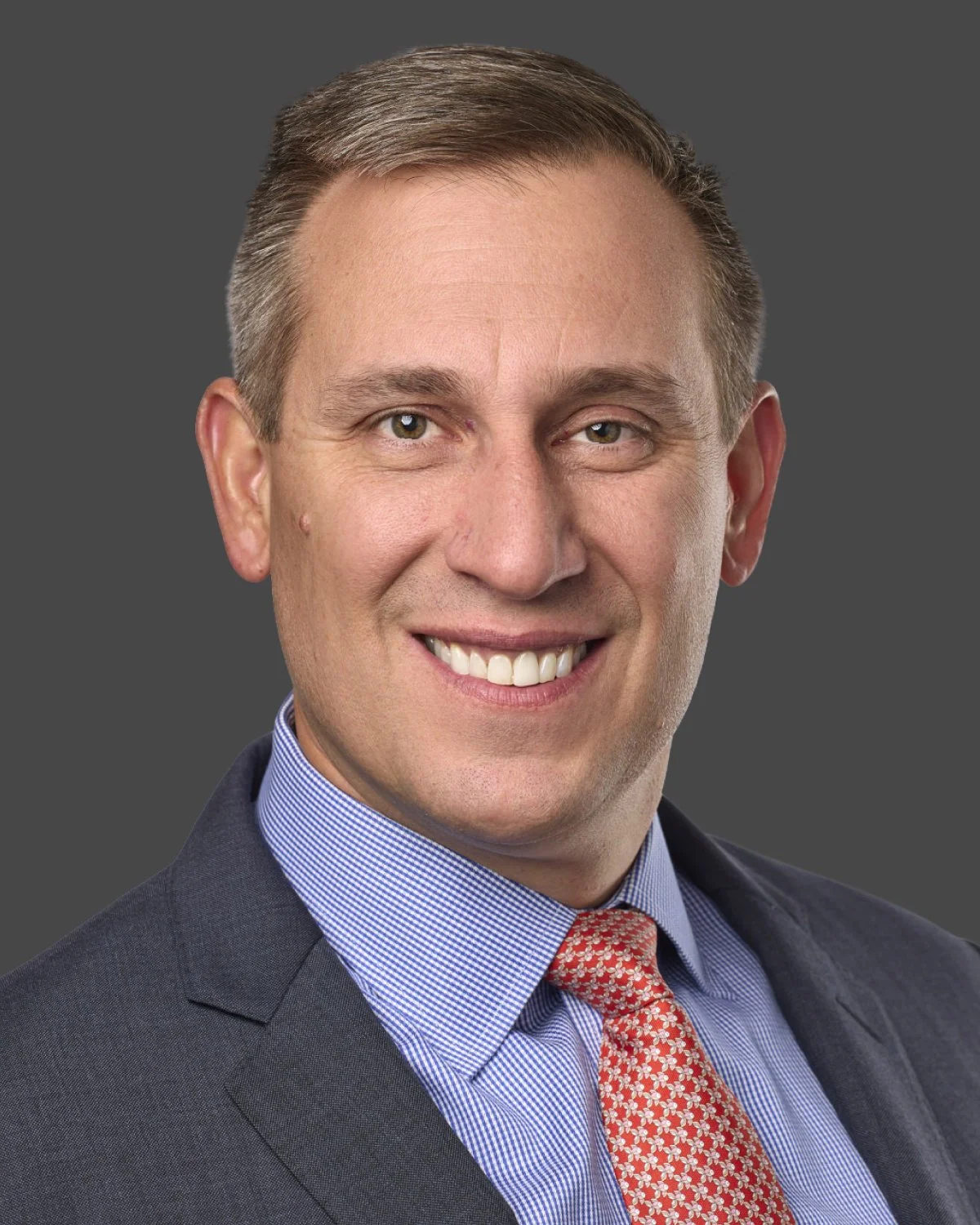 A smiling man in a business suit and red patterned tie against a gray background.