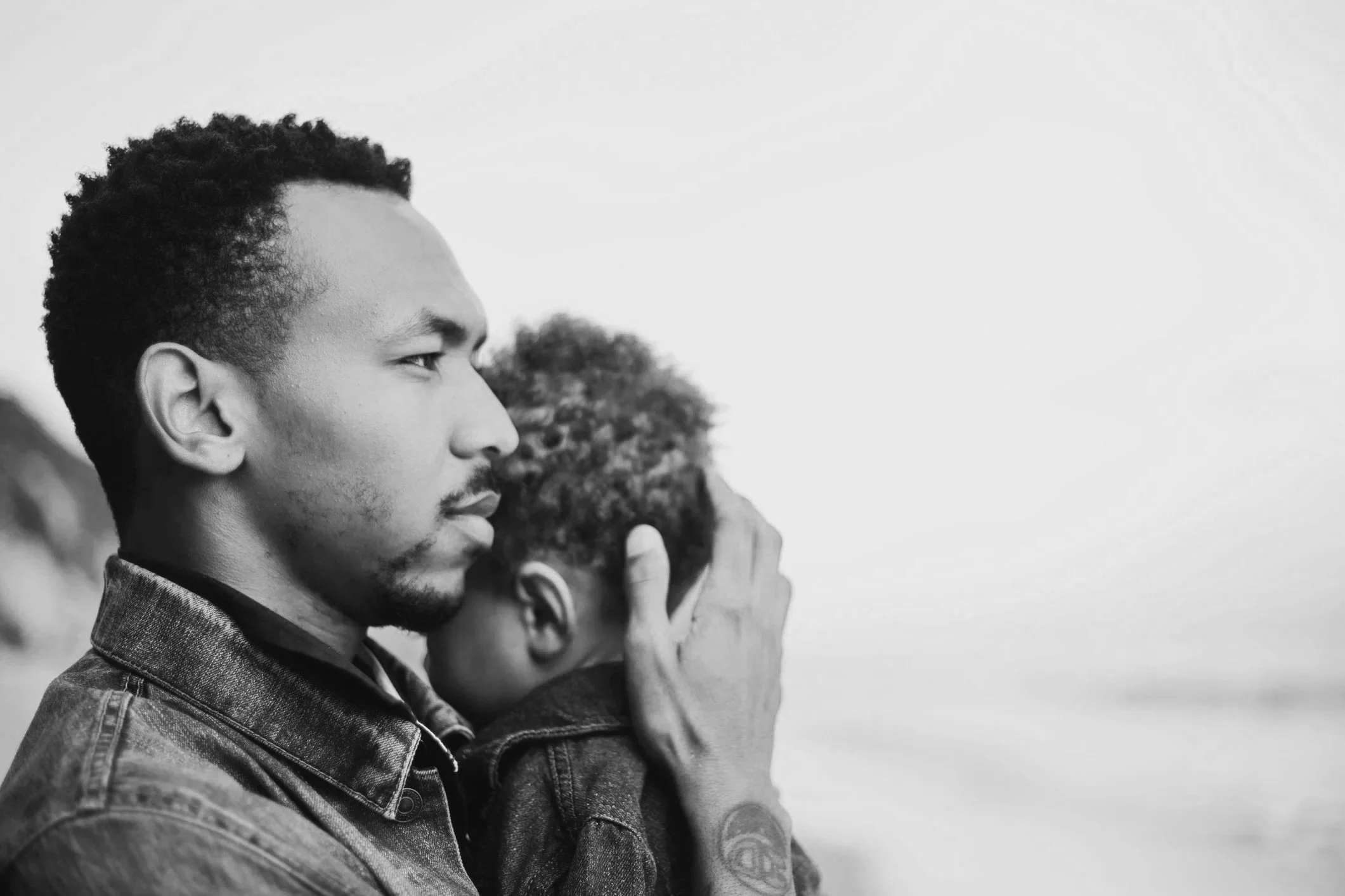 Close-up of a young man with curly hair, beard, and wearing a denim jacket, with others in the background, during a moment of reflection or prayer.