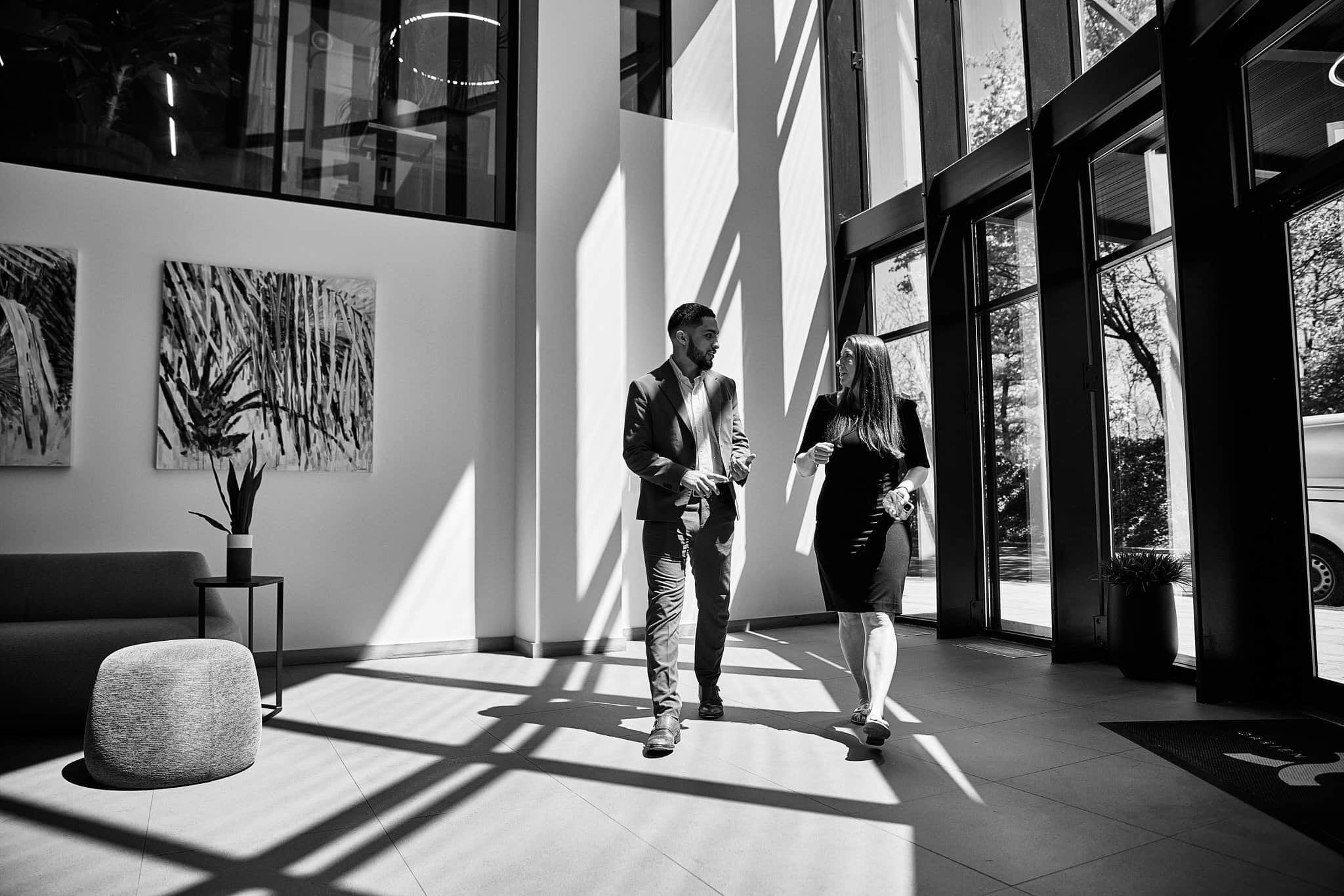 Two people, a man and a woman, are standing and talking inside a modern building with large glass windows that cast shadows on the floor and wall. The interior includes a piece of artwork on the wall, a plant, and a sofa.
