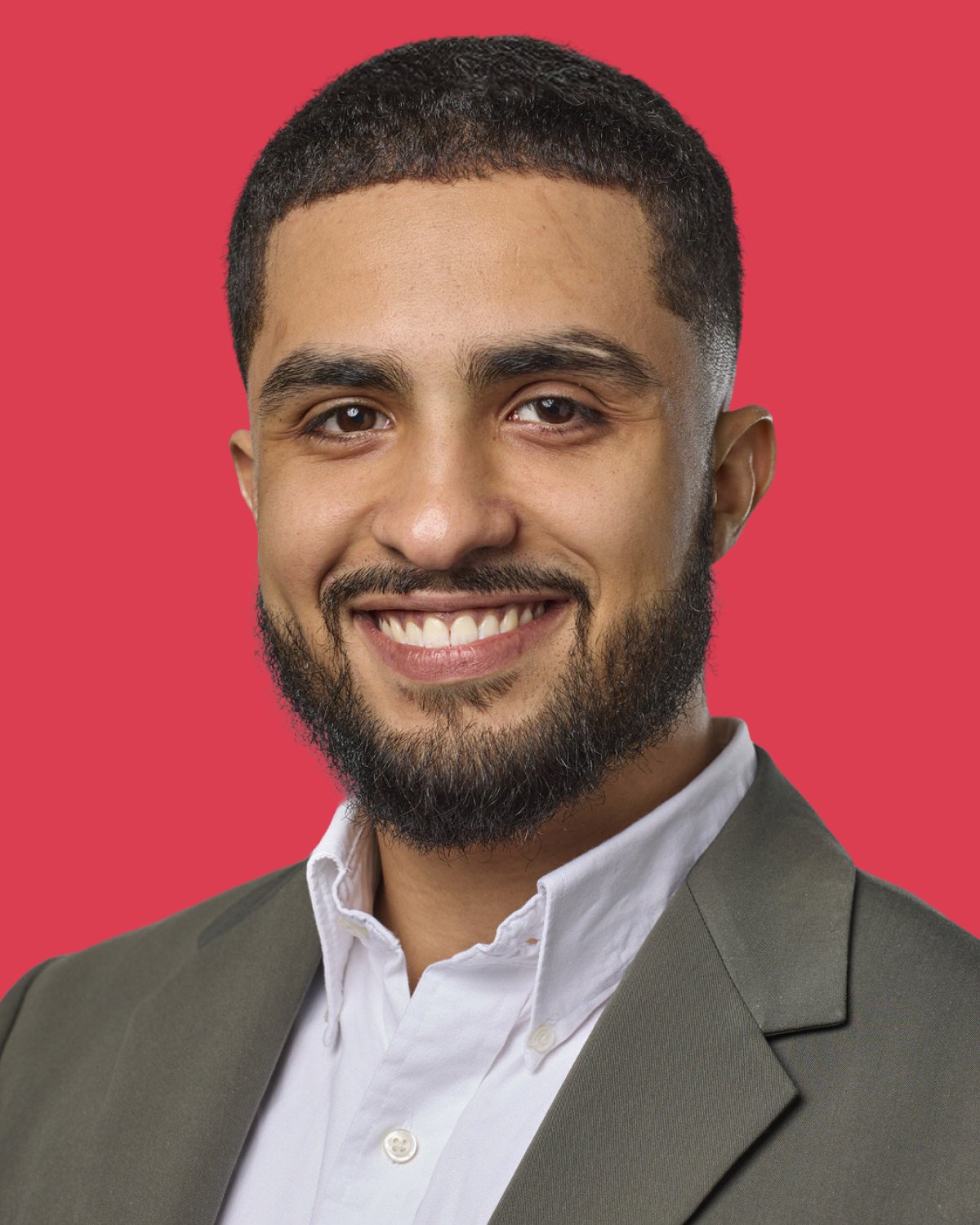 A smiling young man with short dark hair, a beard, wearing a white shirt and a dark green blazer, standing against a red background.