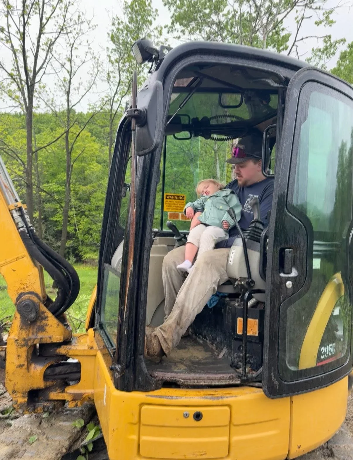 A man sitting in a yellow and black construction vehicle cab holding a sleepy child on his lap. The background shows trees with green leaves.