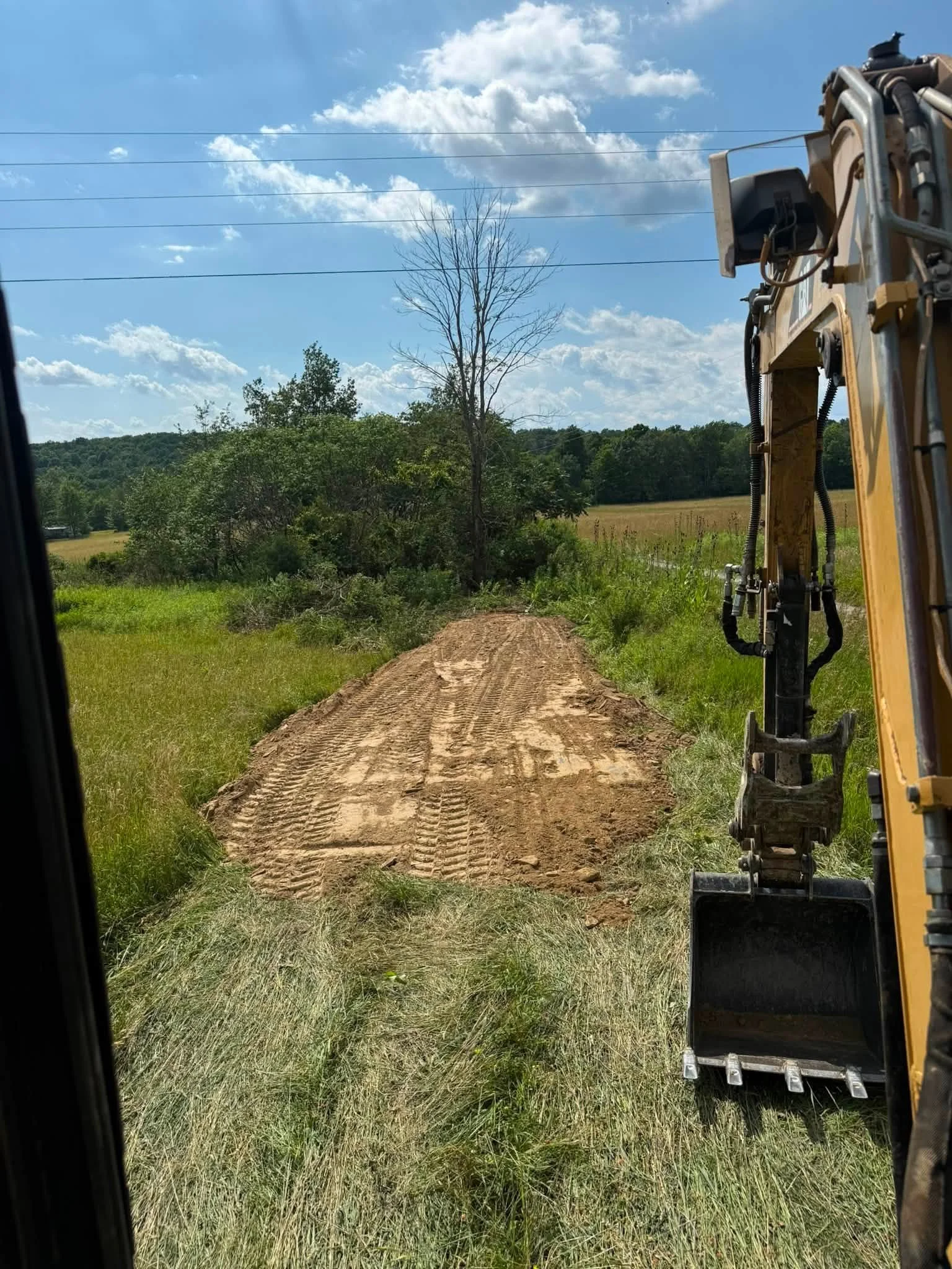 View from a construction vehicle looking down a dirt path through grass and trees on a sunny day with scattered clouds.