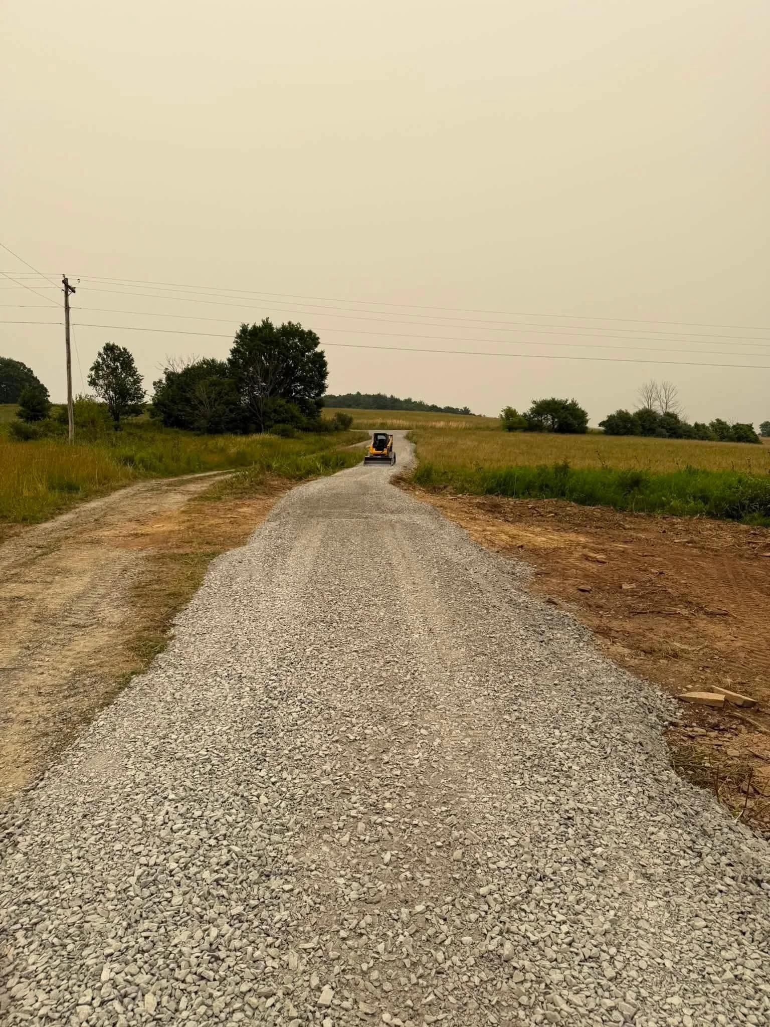 A gravel country road extending into the distance with a small bulldozer on it. The scene is surrounded by grassy fields and scattered trees under a overcast sky.