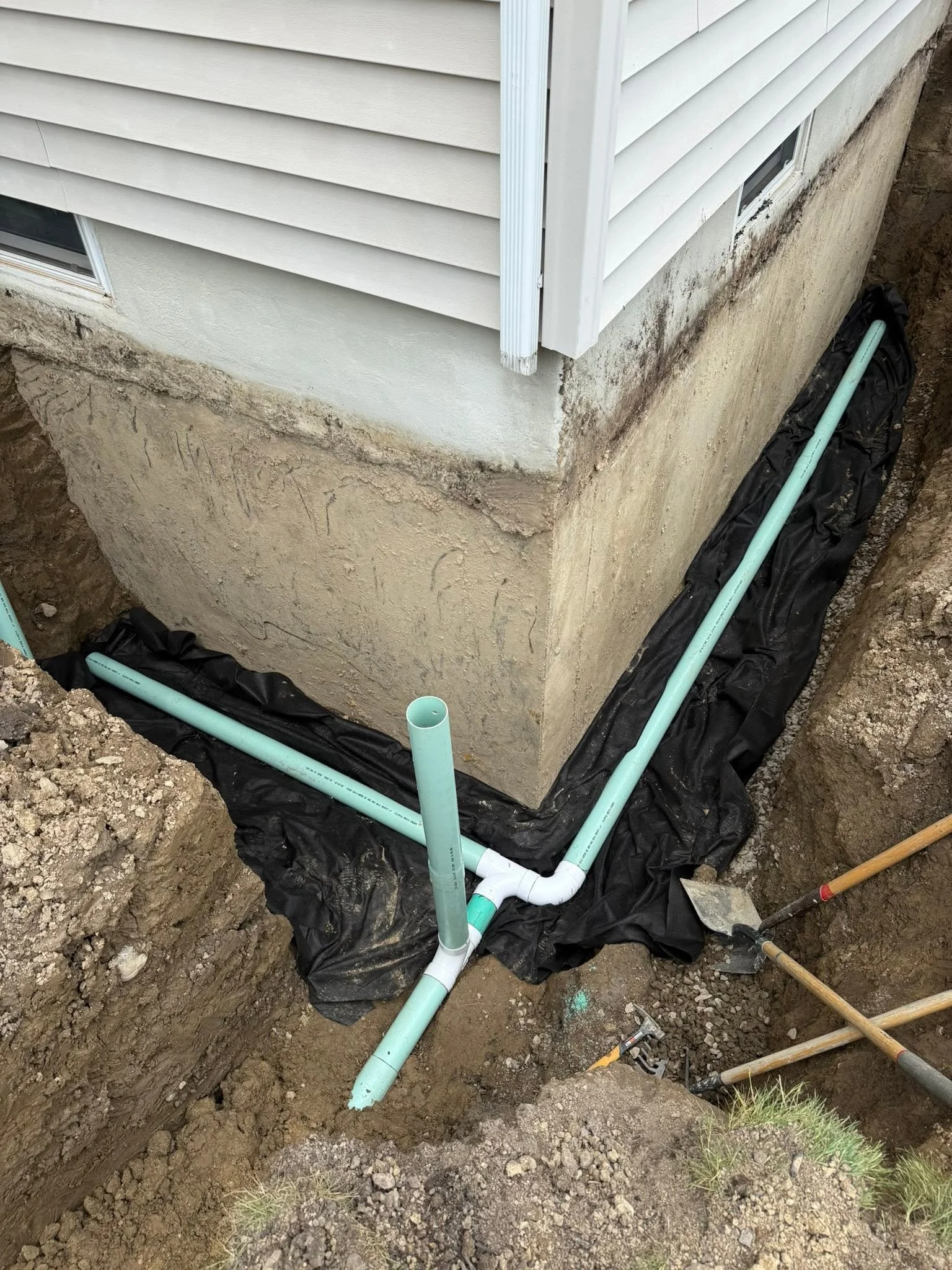 Underground plumbing pipes being installed outside a house near a foundation, with black drainage fabric laid around the pipes.