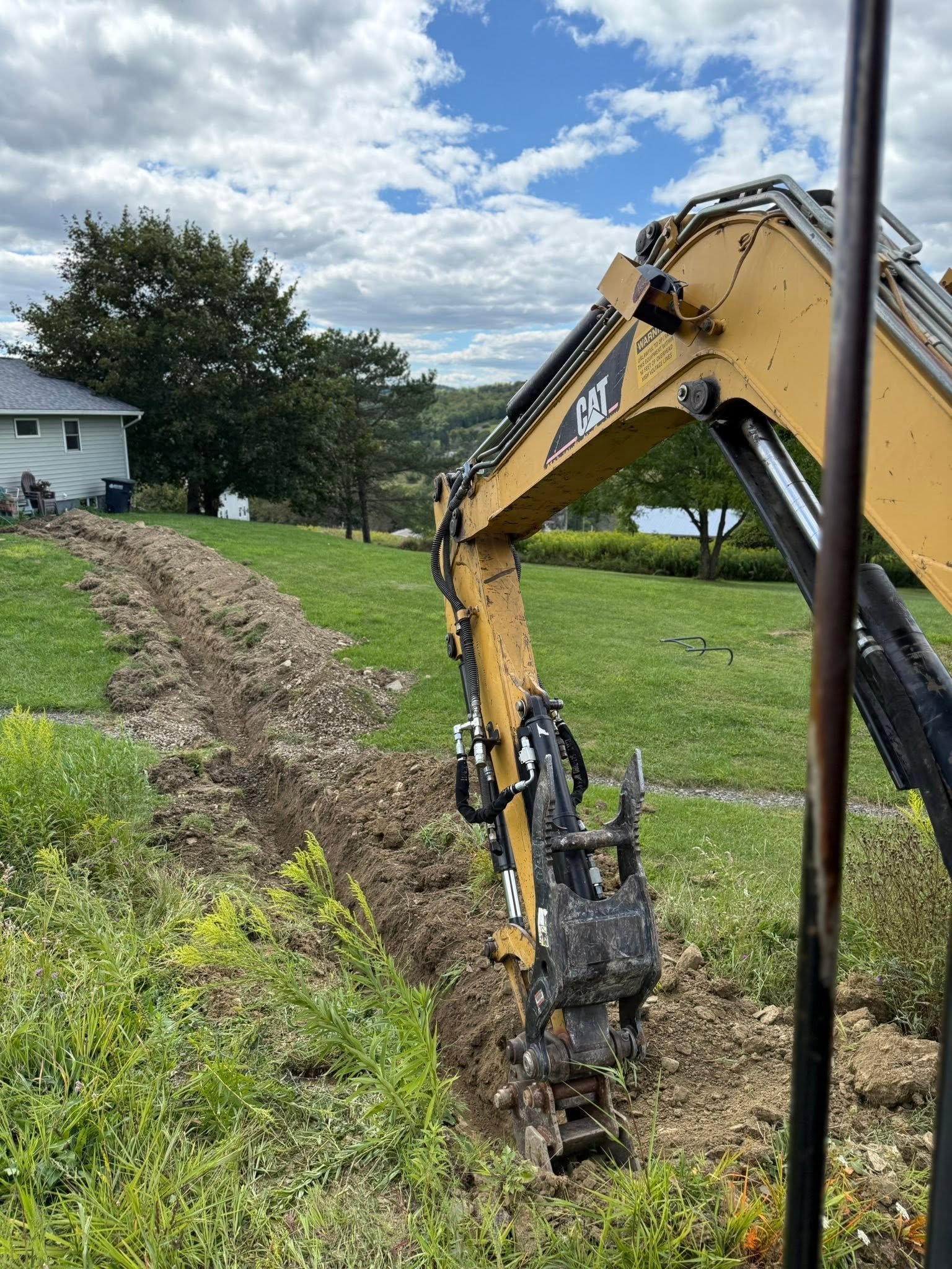 A yellow Caterpillar excavator digging a trench in a grassy yard, with trees, a house, and a cloudy sky in the background.