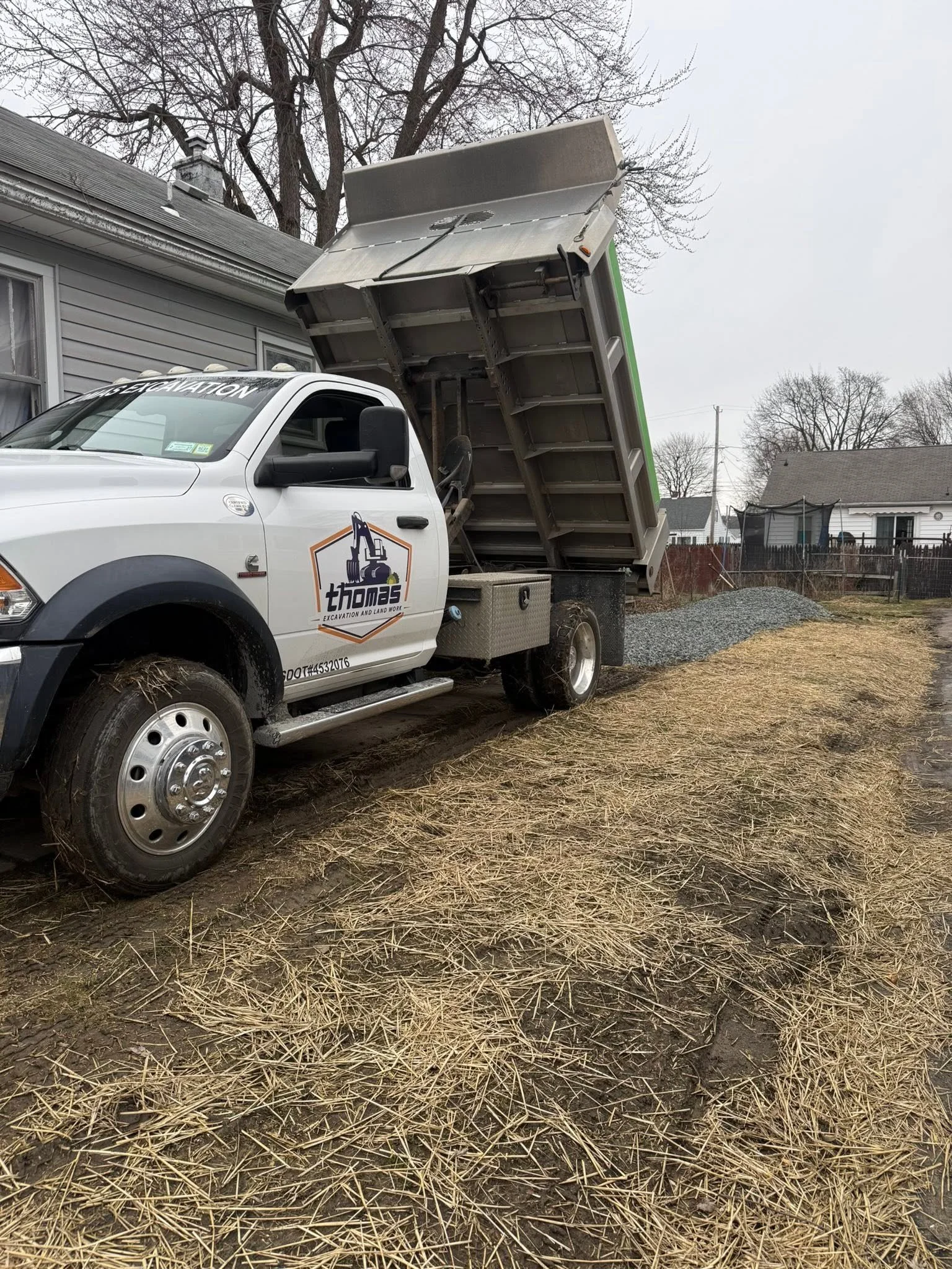 A white dump truck with the word 'Thomes Excavation and Land Work' on the side, parked on a patch of dirt and dried grass, with an open bed lifted. There is a house in the background with a cloudy sky and leafless trees.
