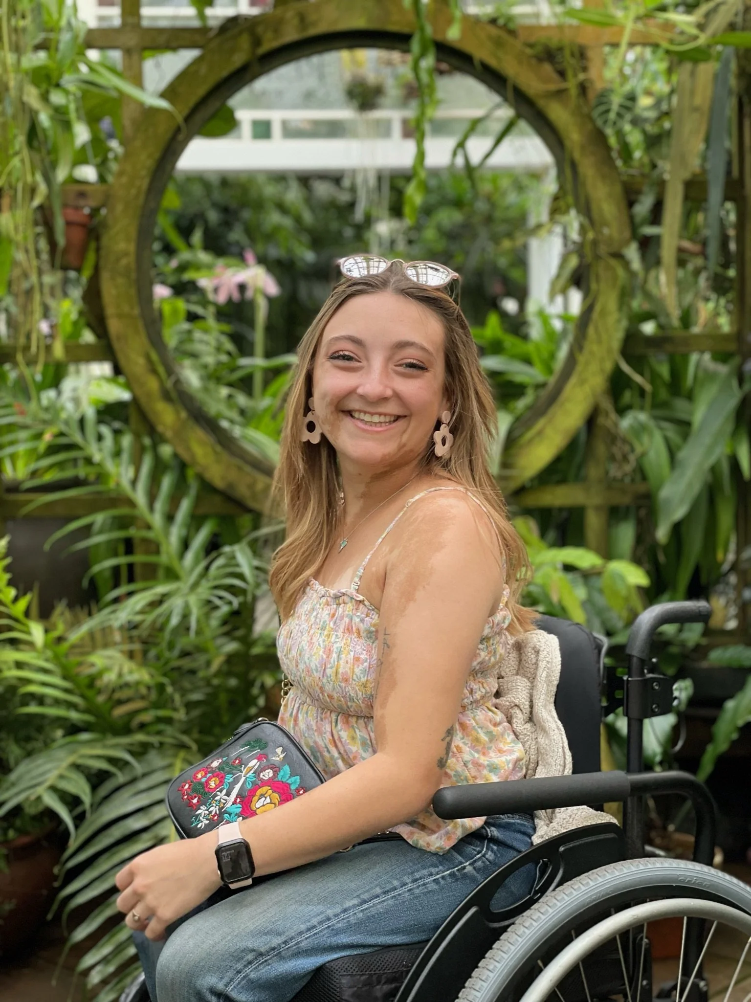 A smiling Sarah Mumper with light brown hair and earrings, sitting in a wheelchair in a lush garden with green plants and wooden structure.