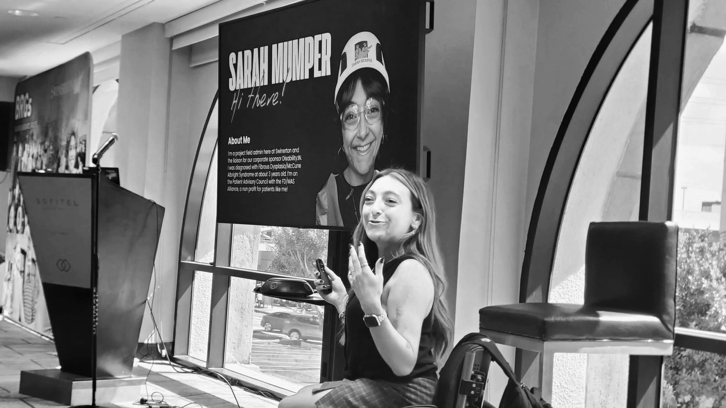 Sarah Mumper sitting in a wheelchair near a large window, with a presentation screen in the background that reads 'Sarah Mumper Hi There!' and an 'About Me' section.