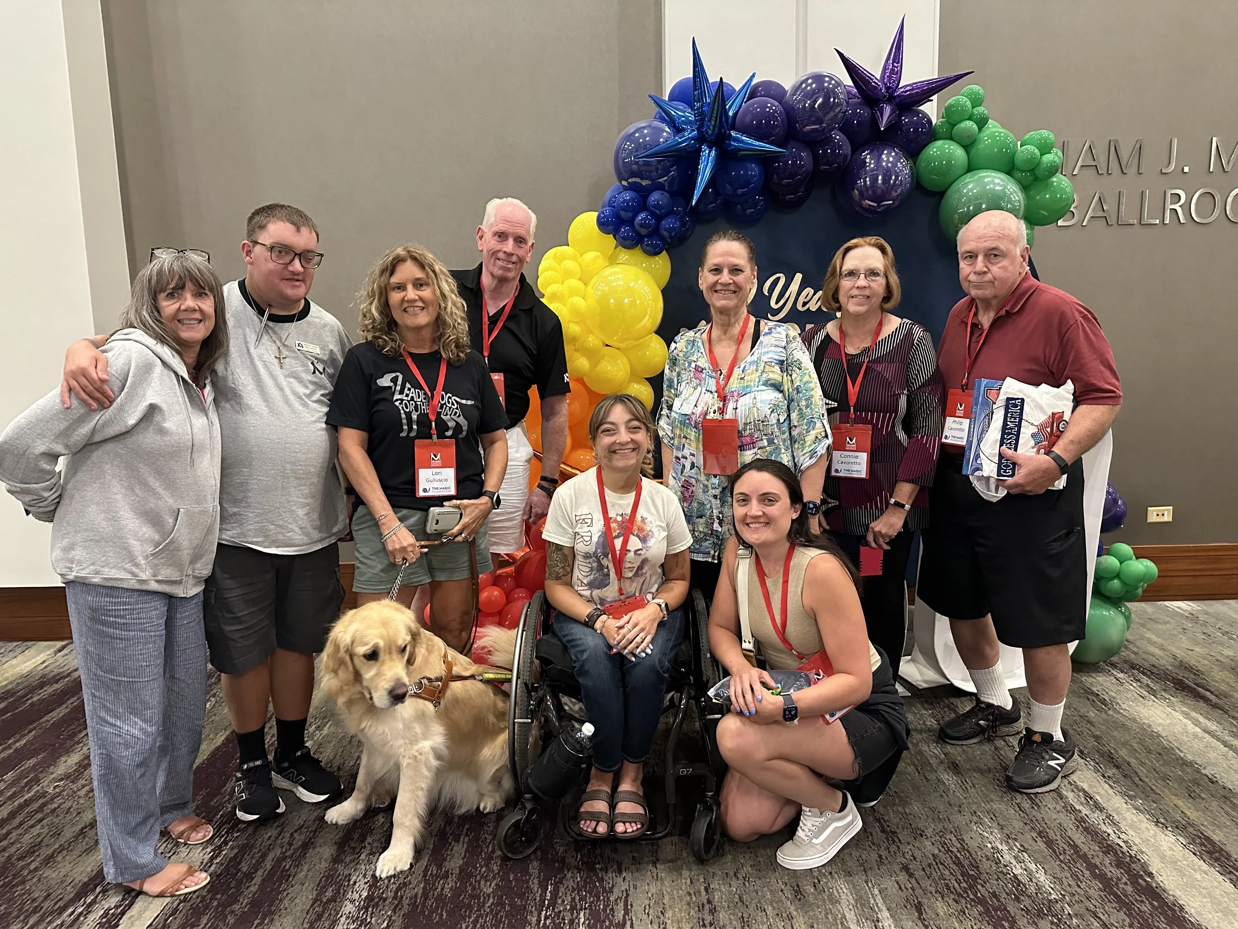 Group of seven people including Sarah in her wheelchair and a guide dog in front of balloon decorations at the MAGIC Foundation, some wearing red lanyards and badges.