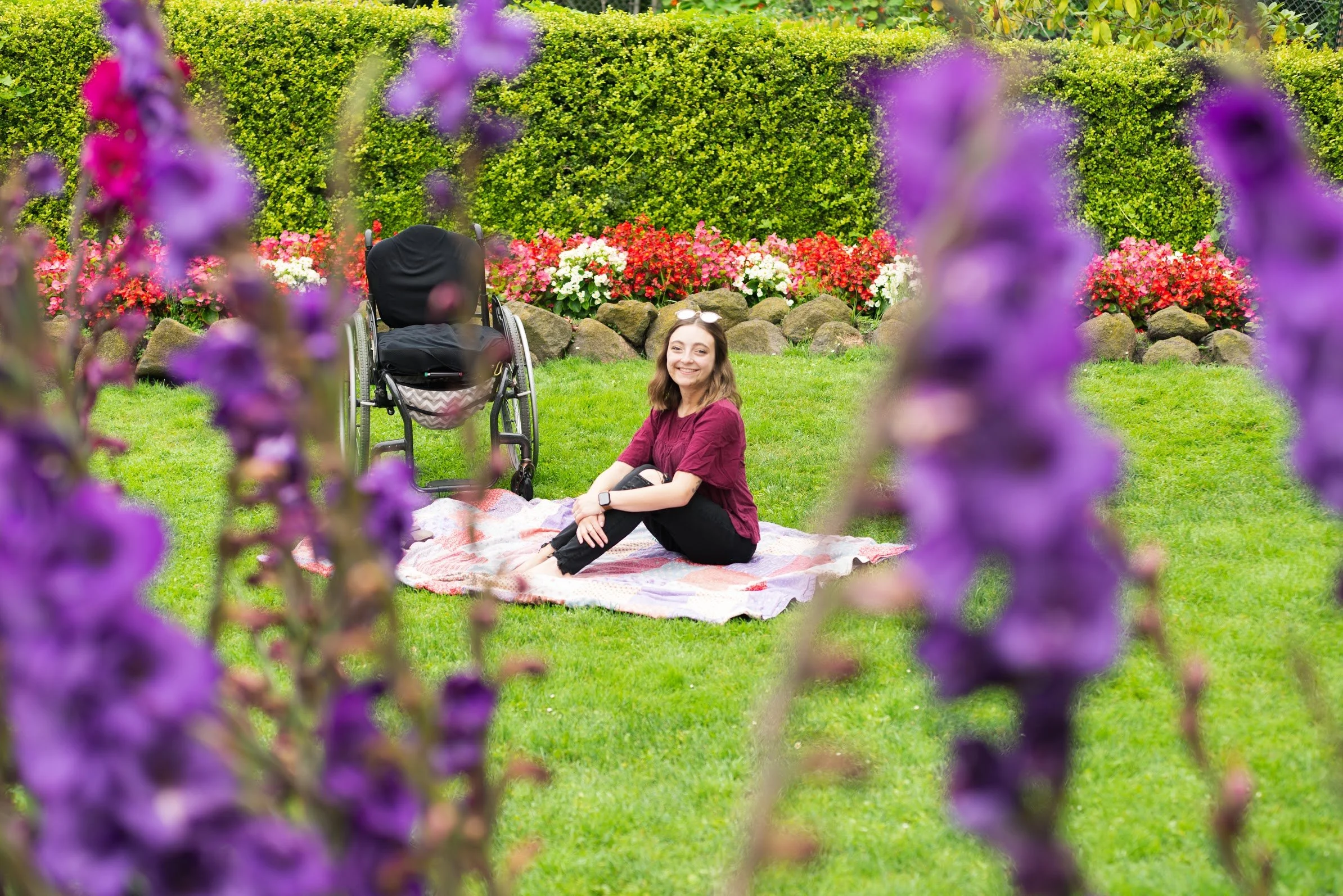 Sarah Mumper sitting on a blanket in a garden with blooming flowers and green grass. In the background, a wheelchair is partially visible. The garden is vibrant with various colorful flowers and trimmed hedges.