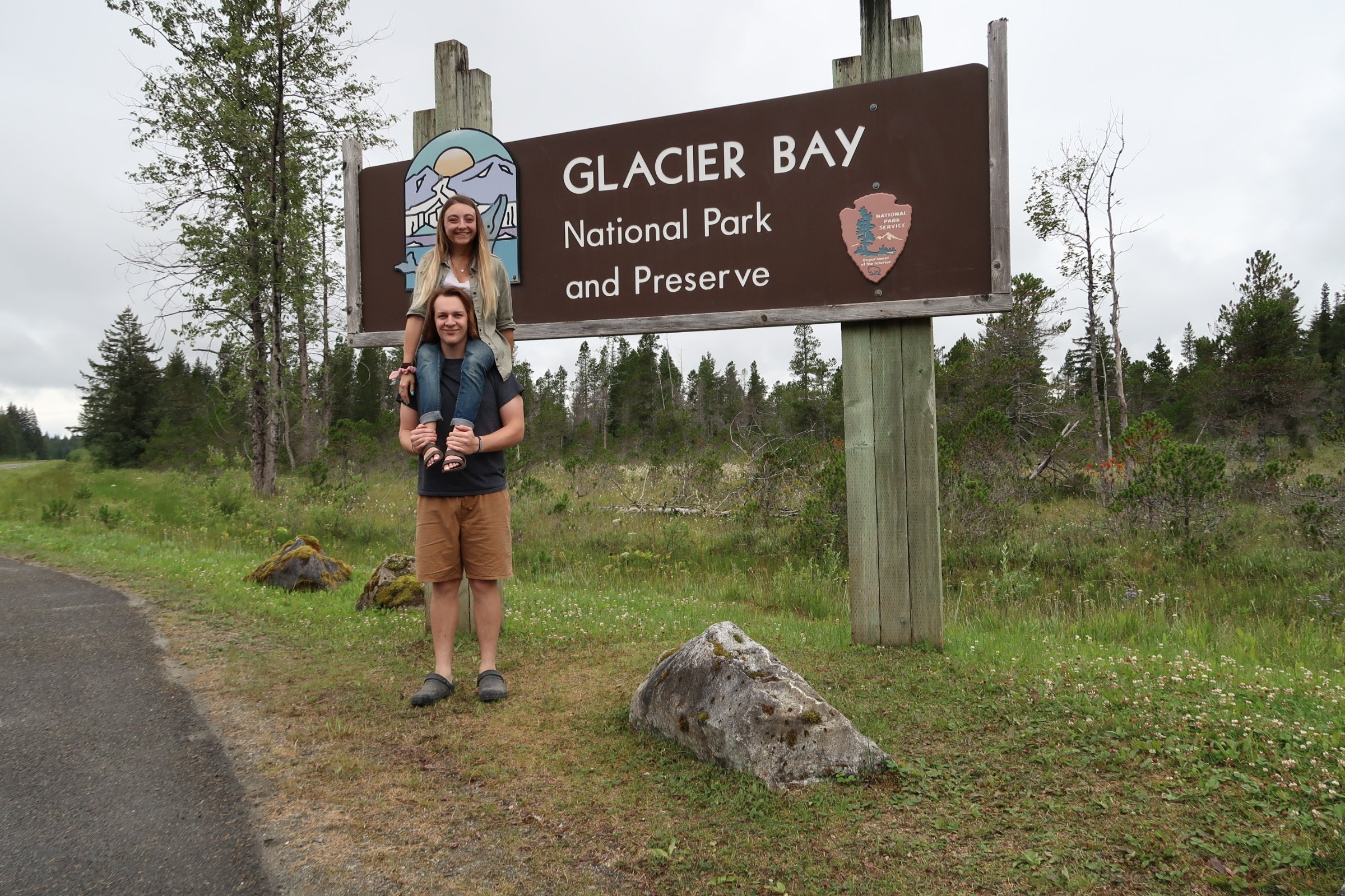 Sarah Mumper and her partner Luke, he is standing and she sitting on his shoulders, smiling in front of a Glacier Bay National Park and Preserve sign surrounded by trees and greenery.