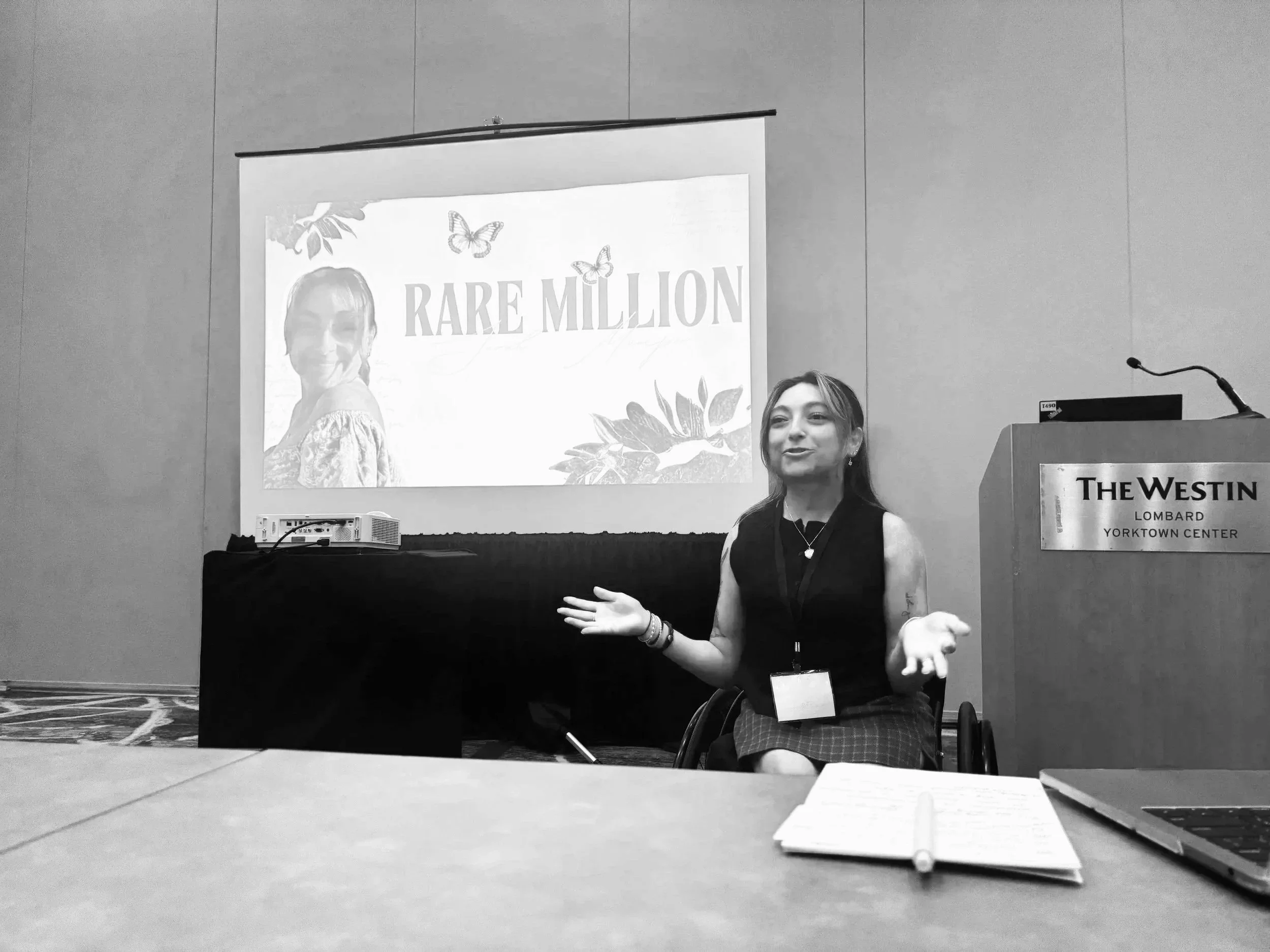 Sarah Mumper sitting in a wheelchair in front of a presentation screen that reads 'Rare Million' with floral and butterfly graphics, at a conference in a hotel conference room.