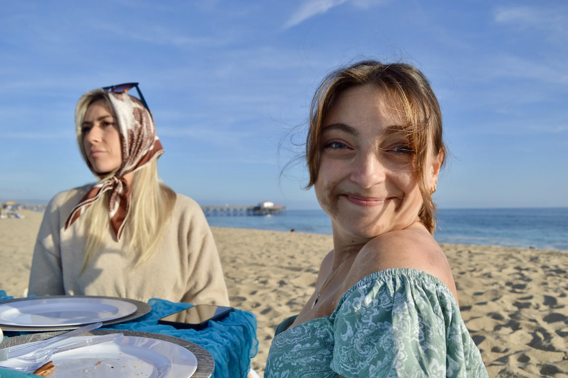 Two women (Sarah with brown hair smiling at the camera and Haley looking off camera wearing a scarf on her head with sunglasses) sit at a beachside picnic table with plates of food, smiling, with a pier and ocean in the background on a sunny day.