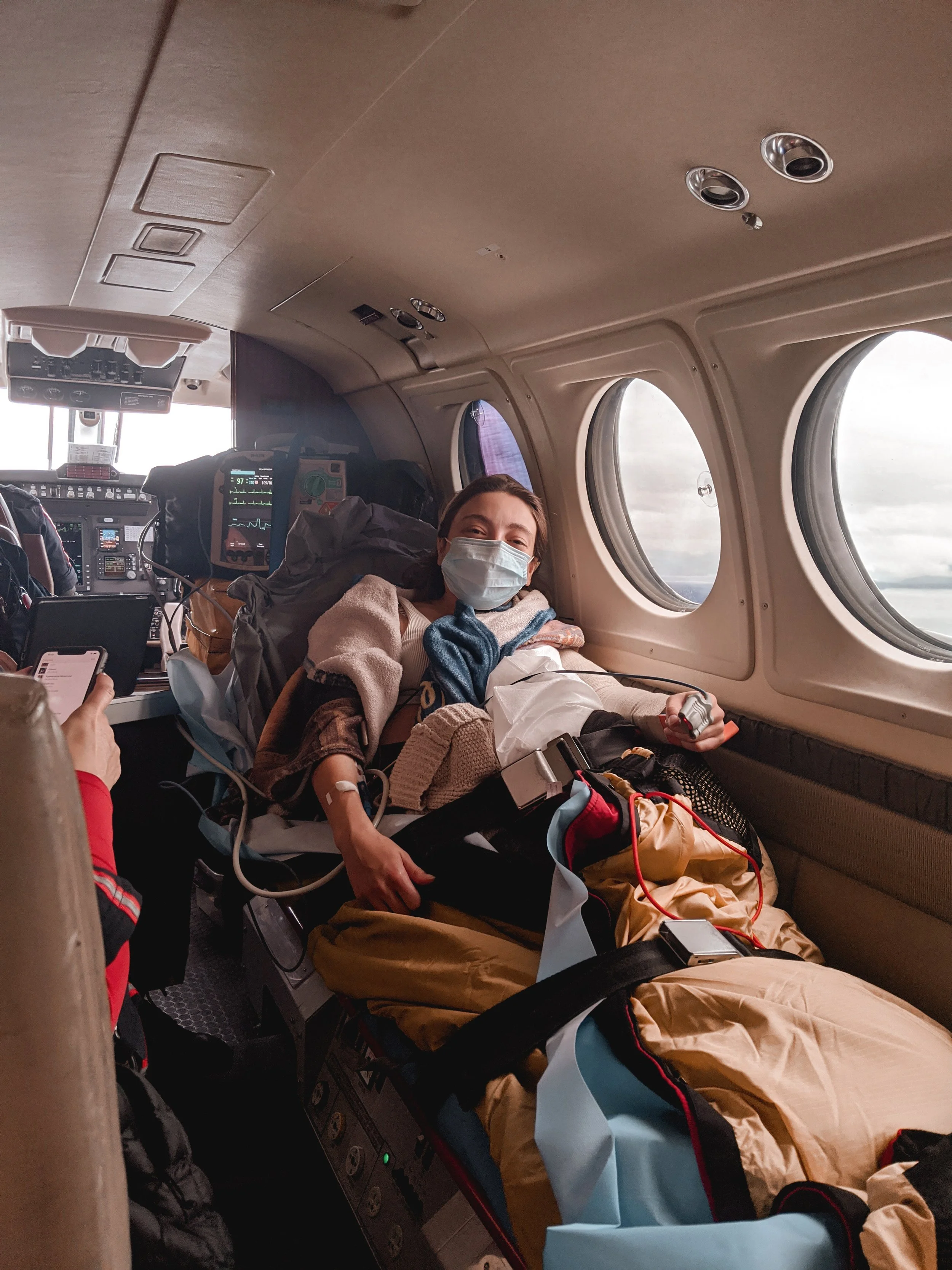 Sarah laying on a gurney, wearing a medical mask, with medical monitors and equipment around her, showing she may be receiving medical treatment during flight for a broken arm.