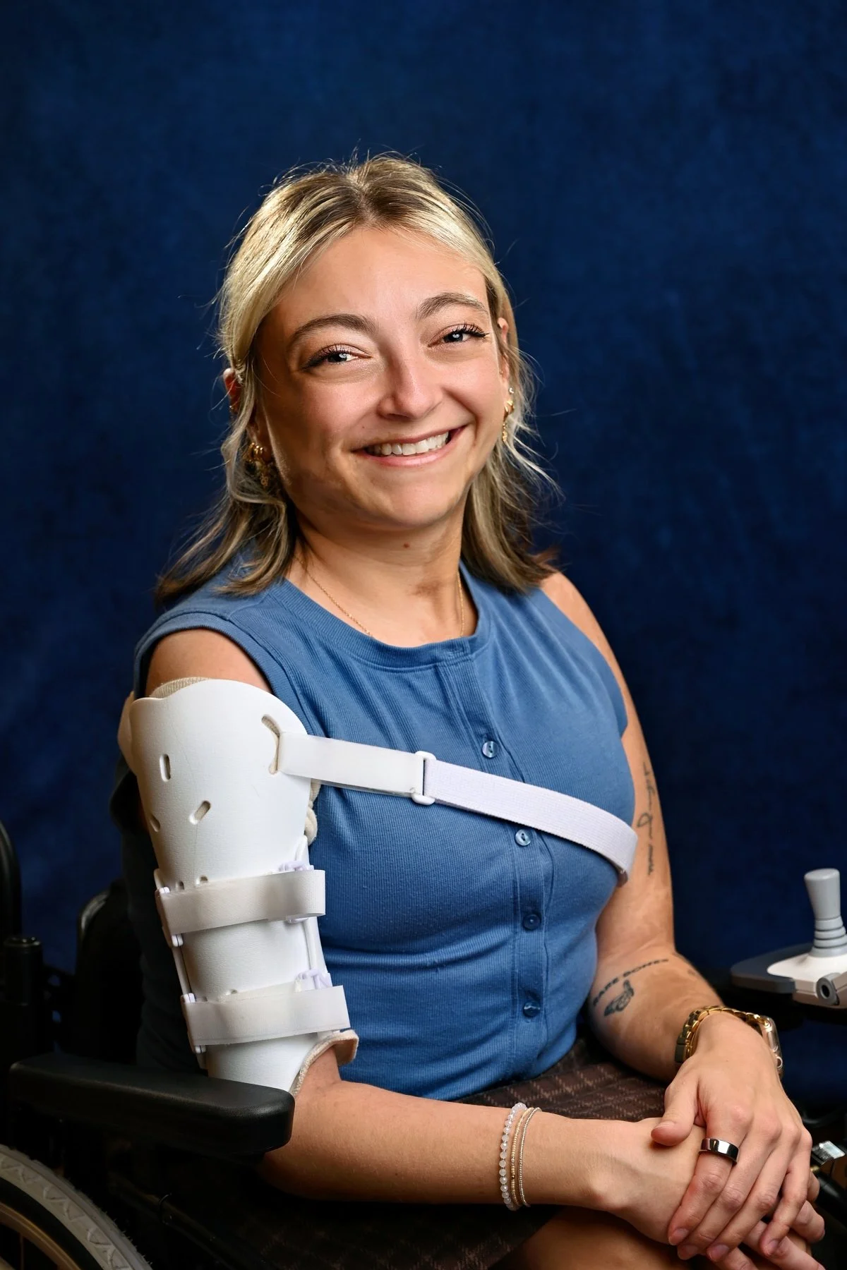 Smiling Sarah with blonde hair in a blue sleeveless top, using a splint on her right humerus, seated in a wheelchair, against a dark blue background.