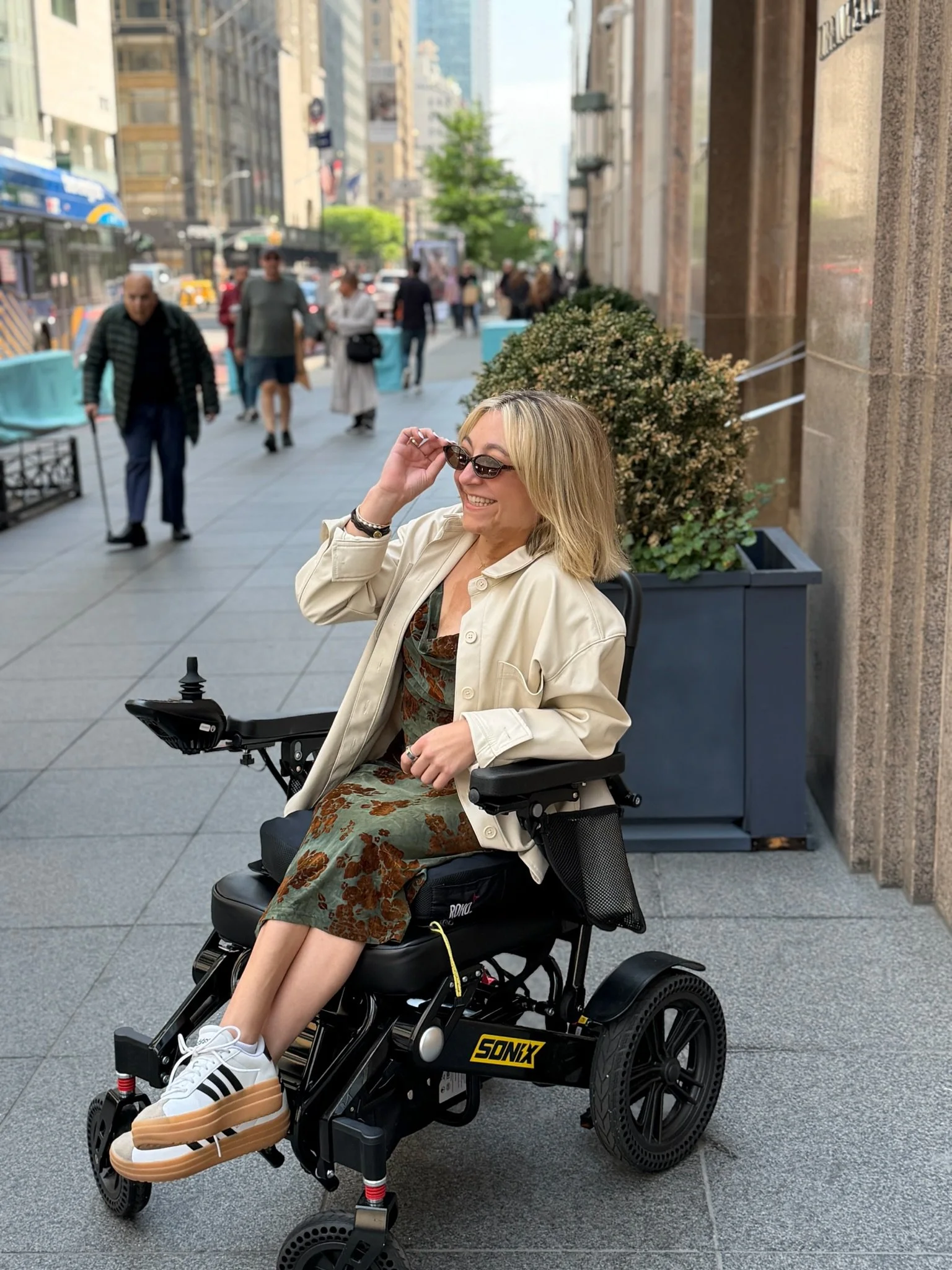 Sarah Mumper smiling and adjusting sunglasses while sitting in a motorized wheelchair on a busy city sidewalk with pedestrians and tall buildings in the background.