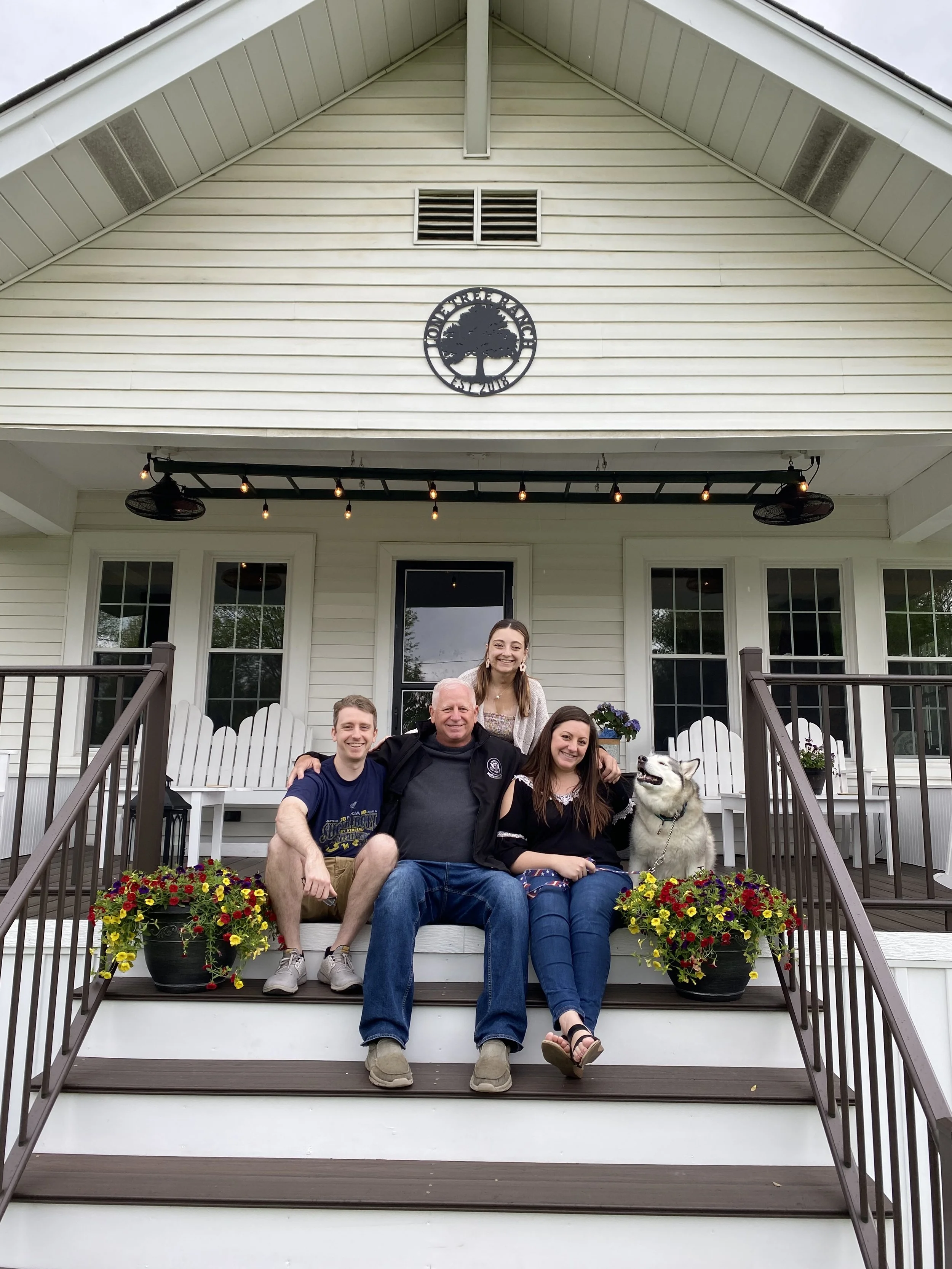 Sarah with her dad, brother, and sister with a husky sitting on a front porch of a house with white siding, surrounded by flower pots with colorful flowers.