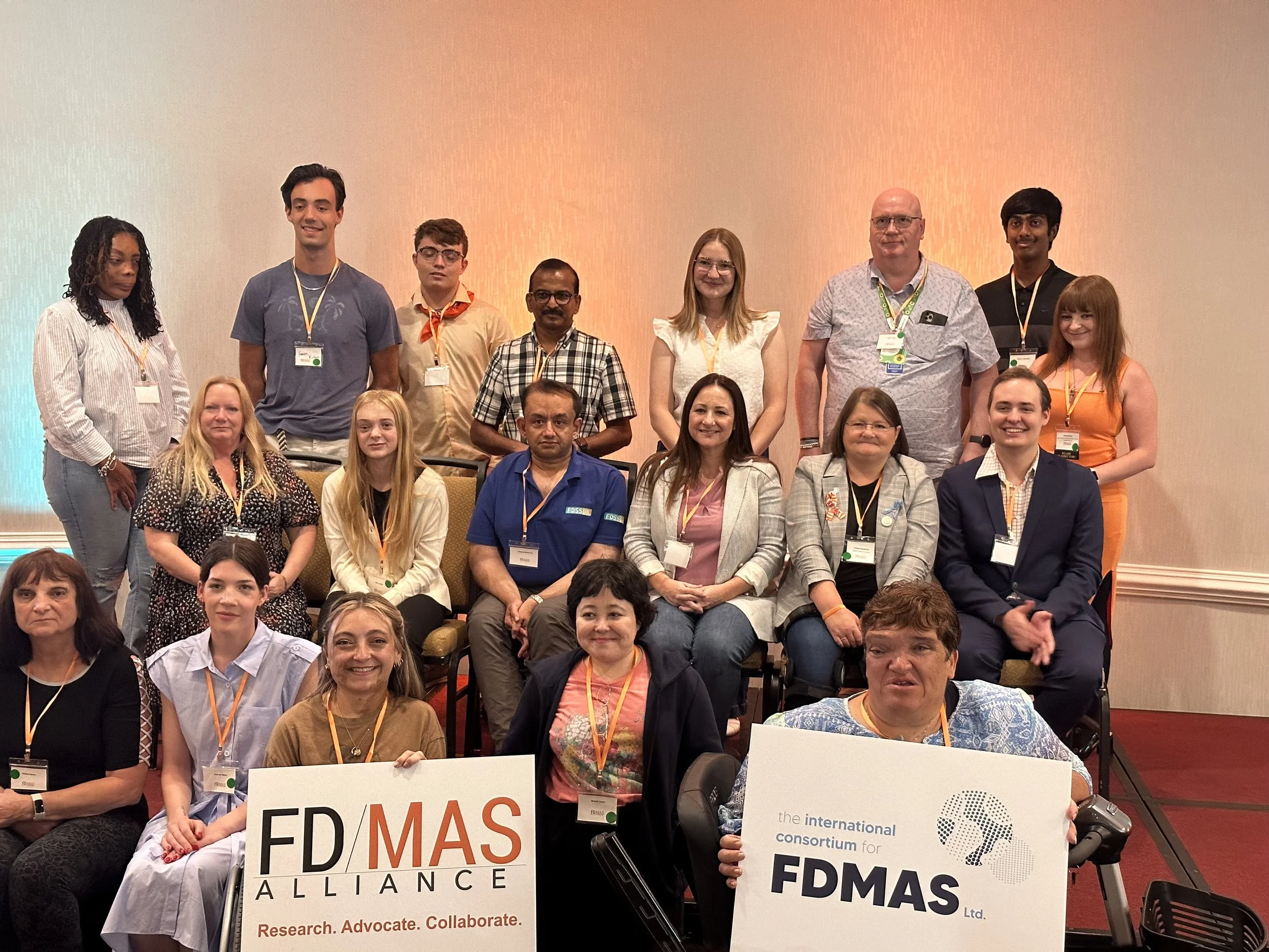 Group of diverse people including Sarah with Fibrous Dysplasia and McCune Albright posing for a photo at an FD/MAS Alliance event, holding signs for the international consortium for FD/MAS and the FD/MAS Alliance, with some wearing conference badges.