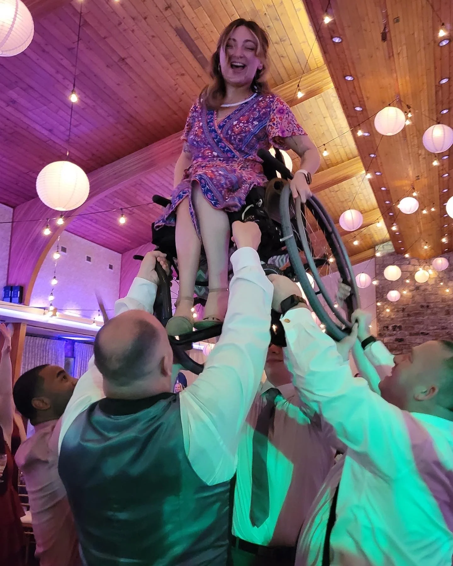 Sarah Mumper in colorful dress being lifted in her wheelchair by several men on a wedding dance floor, with a wooden ceiling and hanging paper lantern lights in the background.