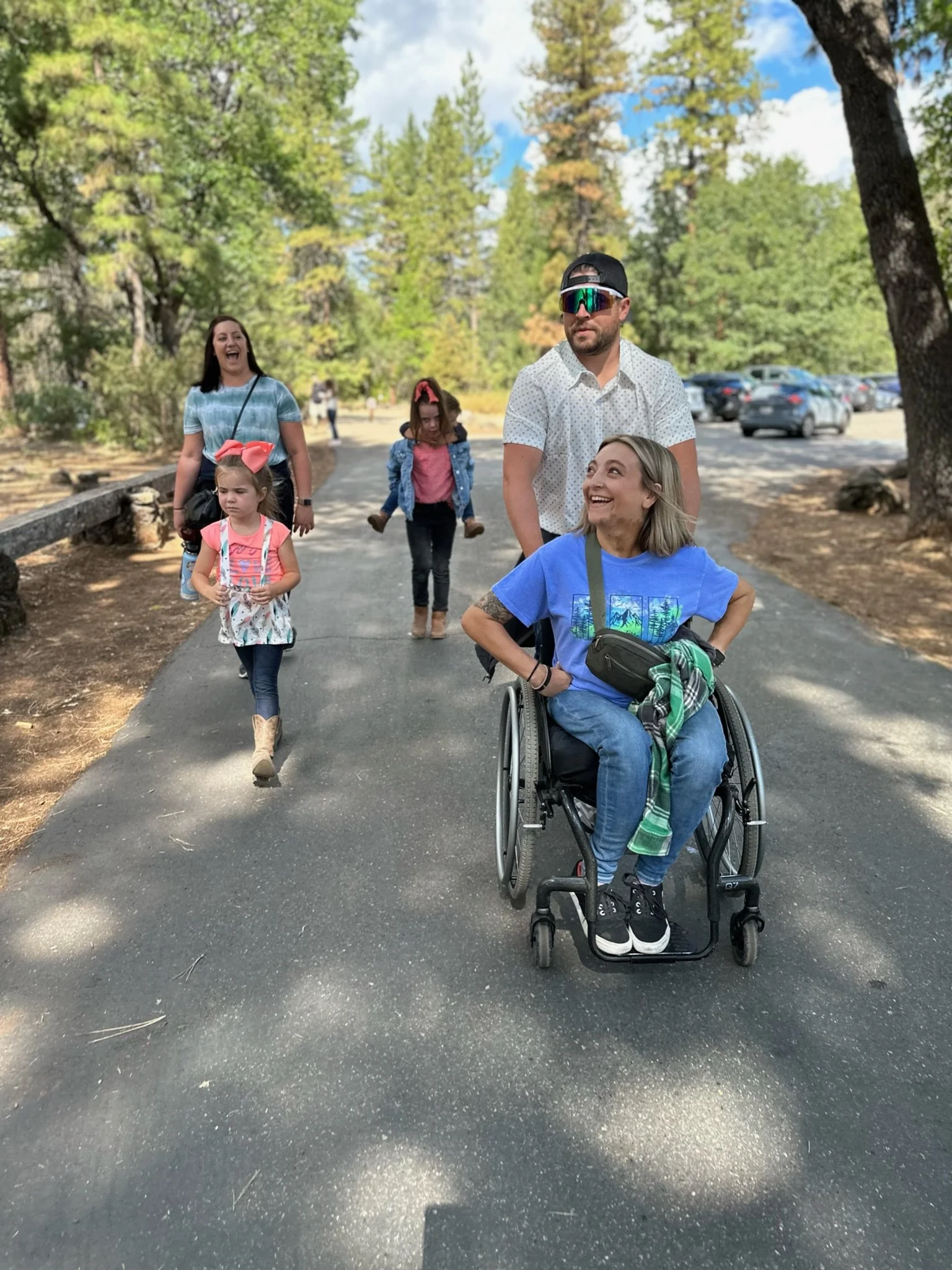 A group of people enjoying a walk outdoors on a paved path in a park surrounded by trees, including a Sarah in a wheelchair smiling, two young girls, and two adults a woman and the man pushing the wheelchair.