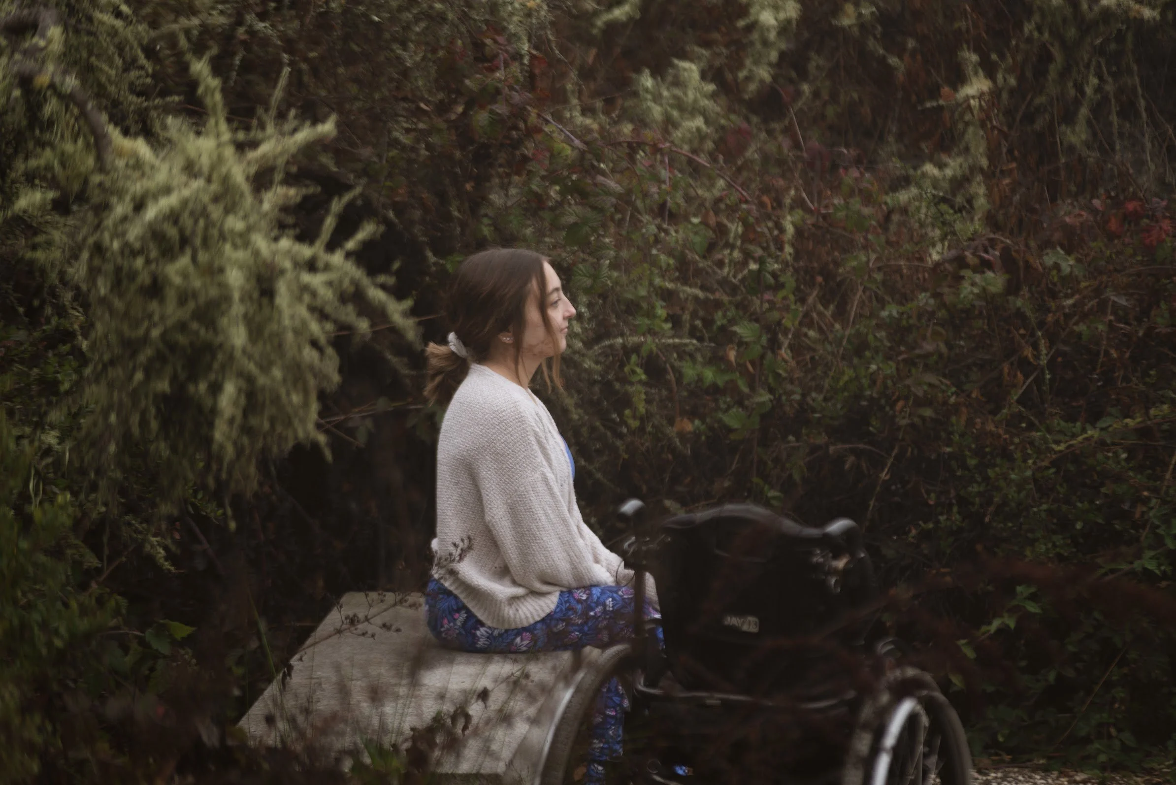 The profile of Sarah Mumper with brown hair tied back sitting on a bench looking out in a natural setting surrounded by greenery and bushes with her wheelchair next to her.
