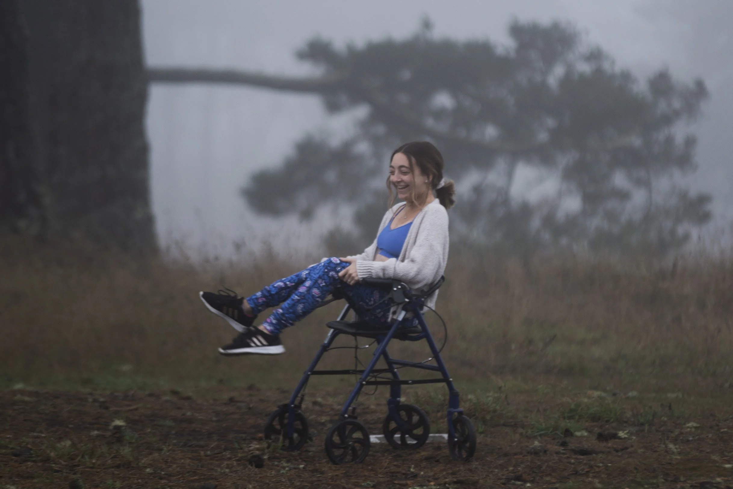 Sarah Mumper with dark hair smiling and sitting on a walker with her legs kicked up outdoors on a cloudy day. She is wearing a blue top, patterned pants, and a gray cardigan. The background features foggy cyprus trees and grass.