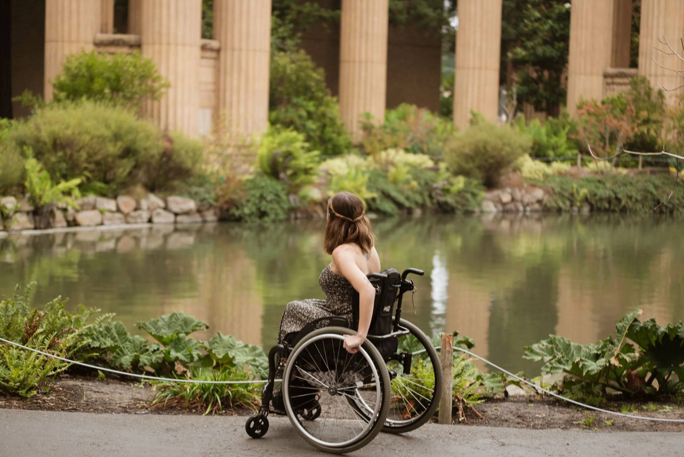 Sarah in a wheelchair sitting by a body of water surrounded by lush greenery with classical columns in the background.