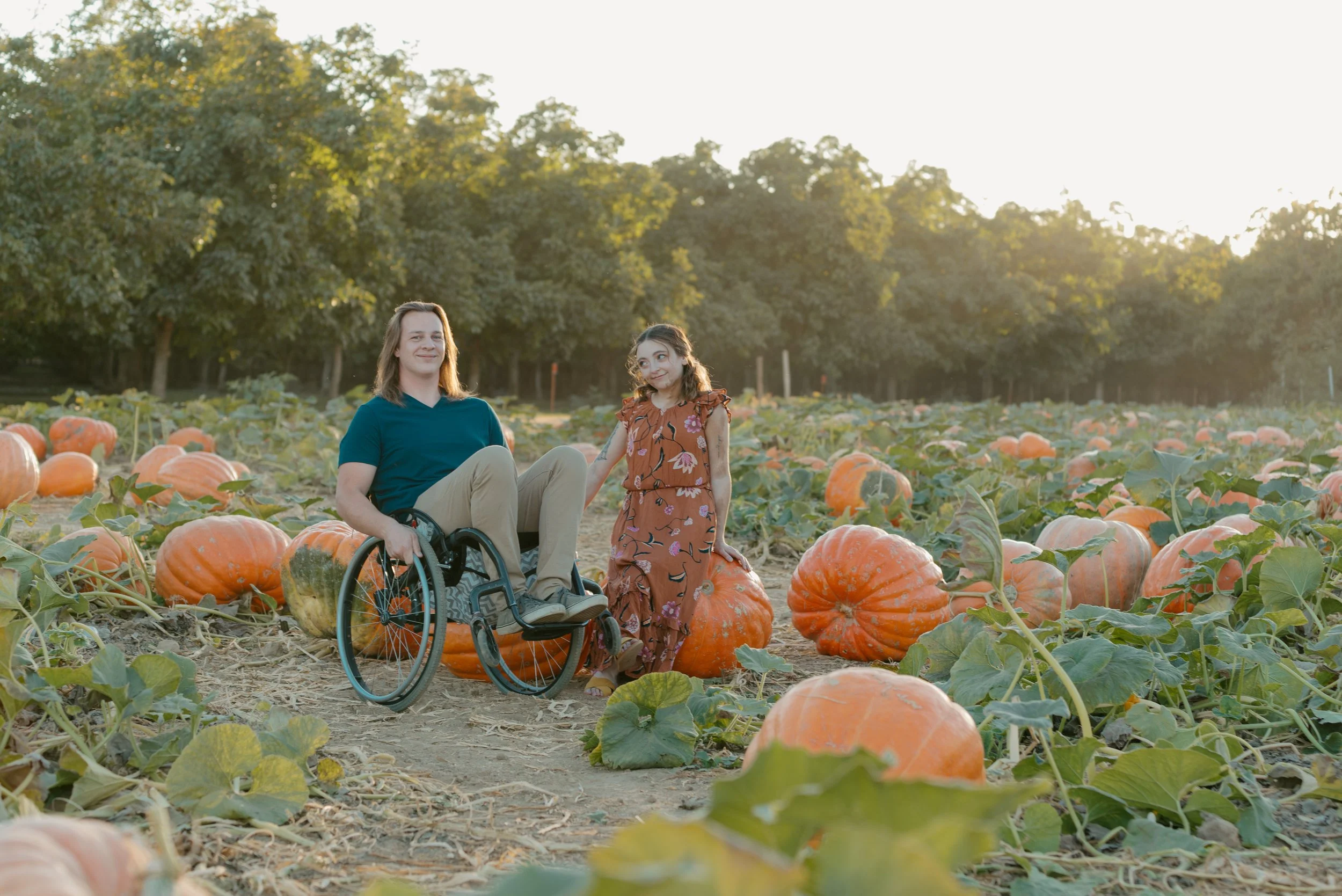 Luke (Sarah's boyfriend) in a wheelchair looking at the camera and Sarah in a patterned dress sitting on large pumpkin in a pumpkin patch, with trees in the background and warm sunlight smirking at Luke.