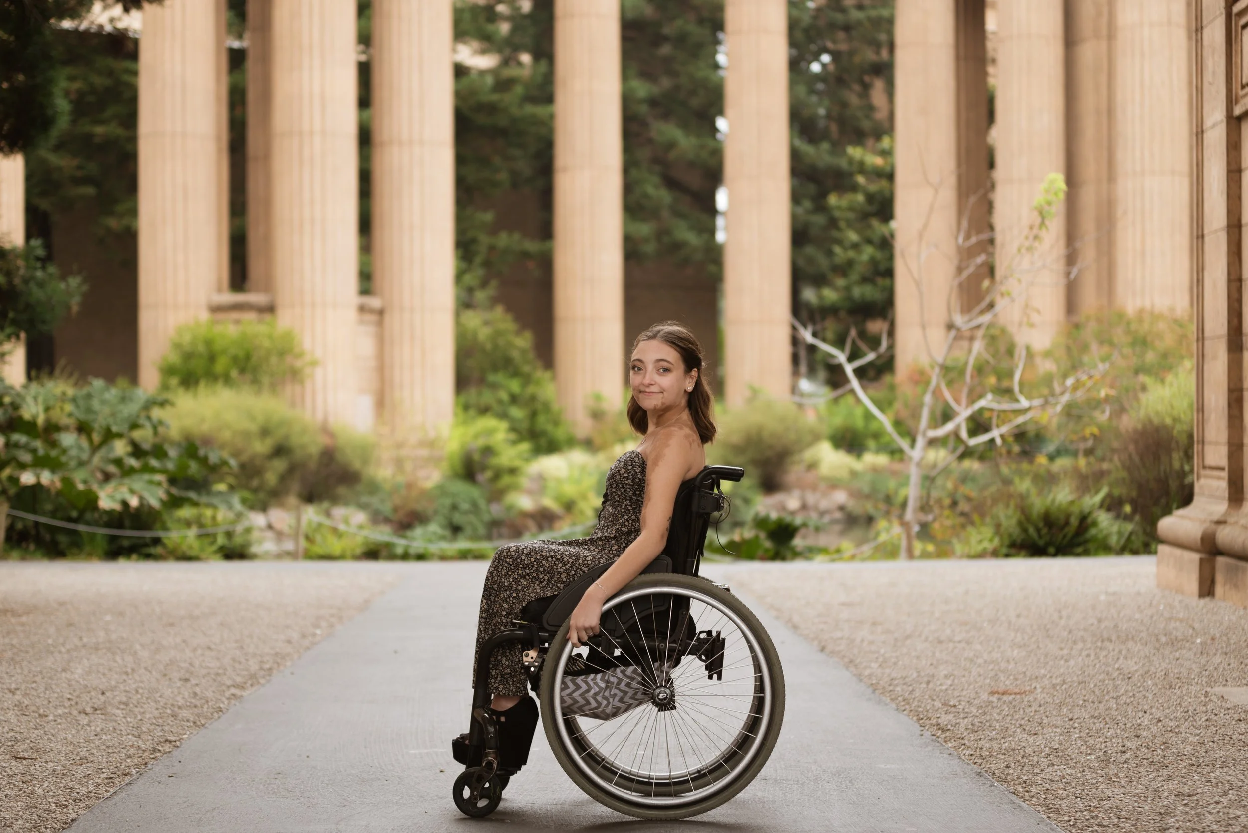 Sarah Mumper in a wheelchair wearing a floral strapless dress sits outdoors in front of ancient Greek-style columns surrounded by greenery.