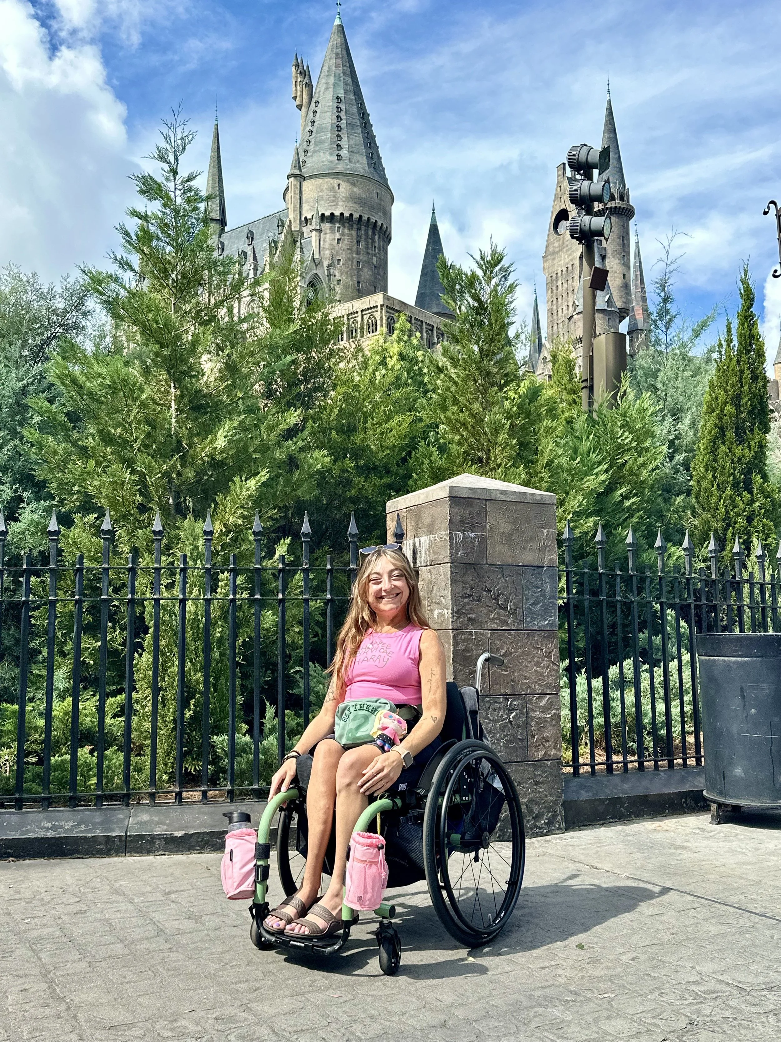 Sarah Mumper in a wheelchair smiling outdoors with the Hogwarts castle in the background and trees surrounding her.