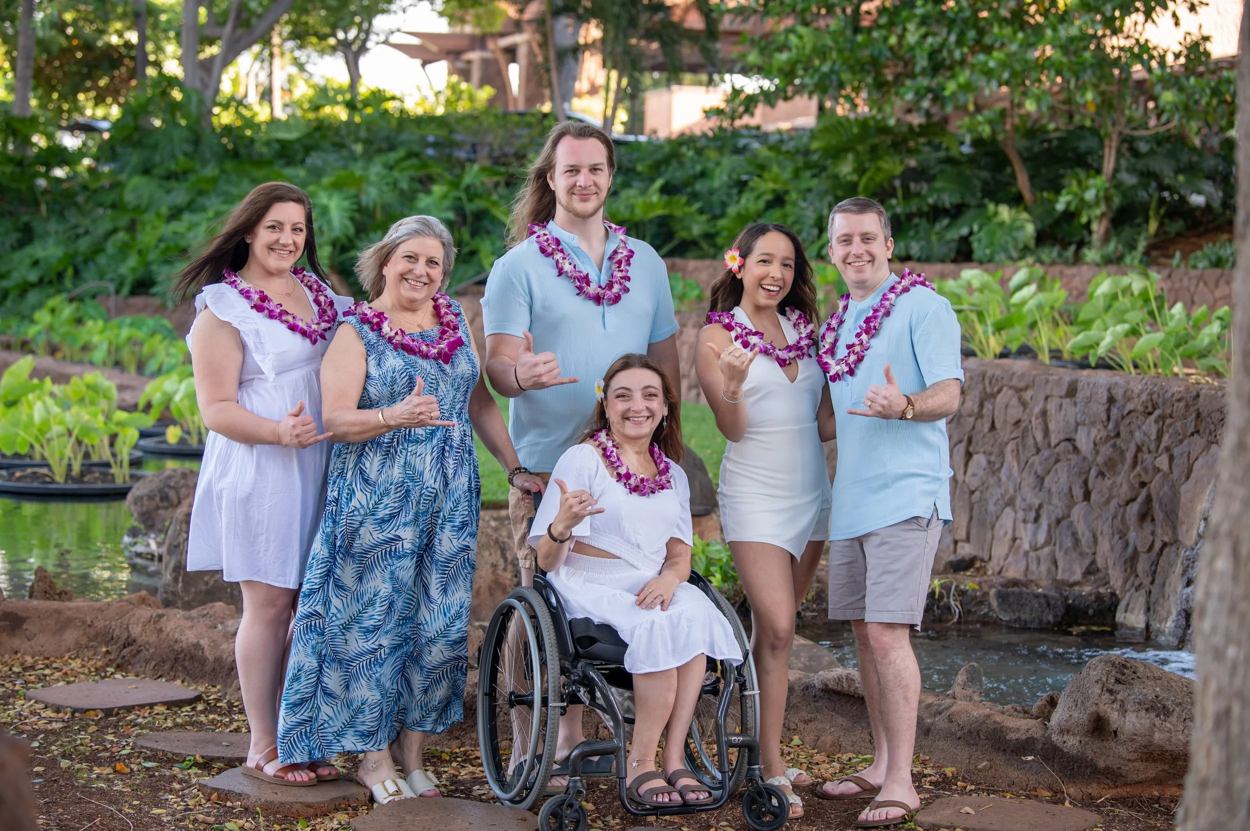Sarah in a wheelchair with her family (mom, sister, brother, sis-in-law, and boyfriend), standing outdoors in Hawaii with lush greenry with a small stream, all wearing leis and smiling at the camera.