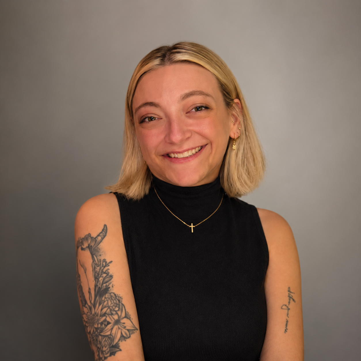 Sarah Mumper with short blonde hair smiling, wearing a black sleeveless turtleneck, gold cross necklace, and earrings, with tattoos on her arms and a gray background.