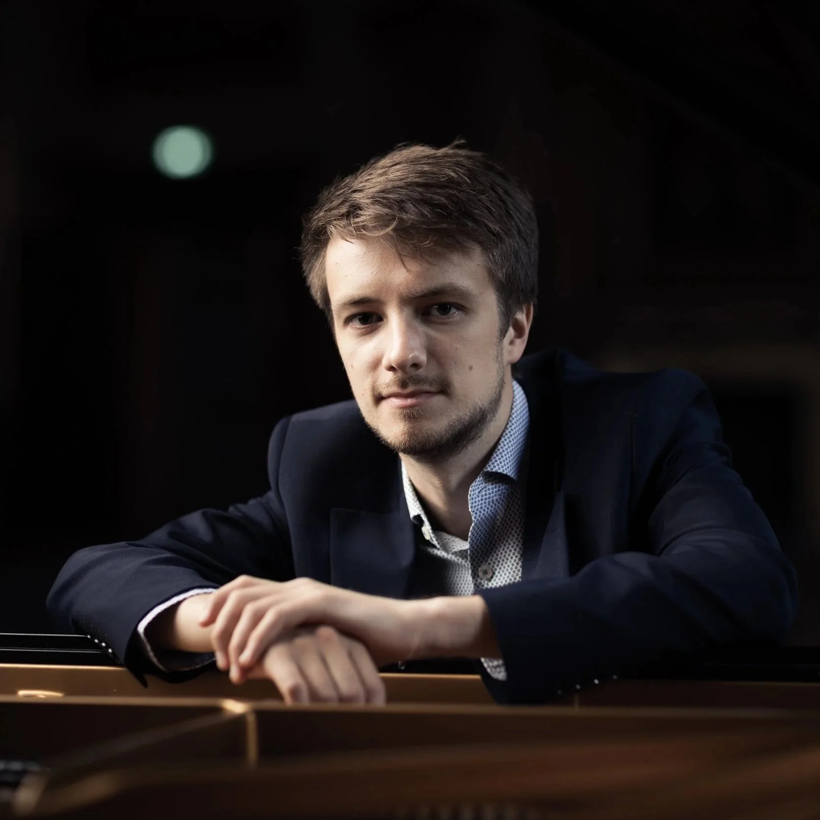Young man with light brown hair, beard, and mustache, wearing a dark suit and light shirt, sitting at a piano with his hands resting on it, against a black background.