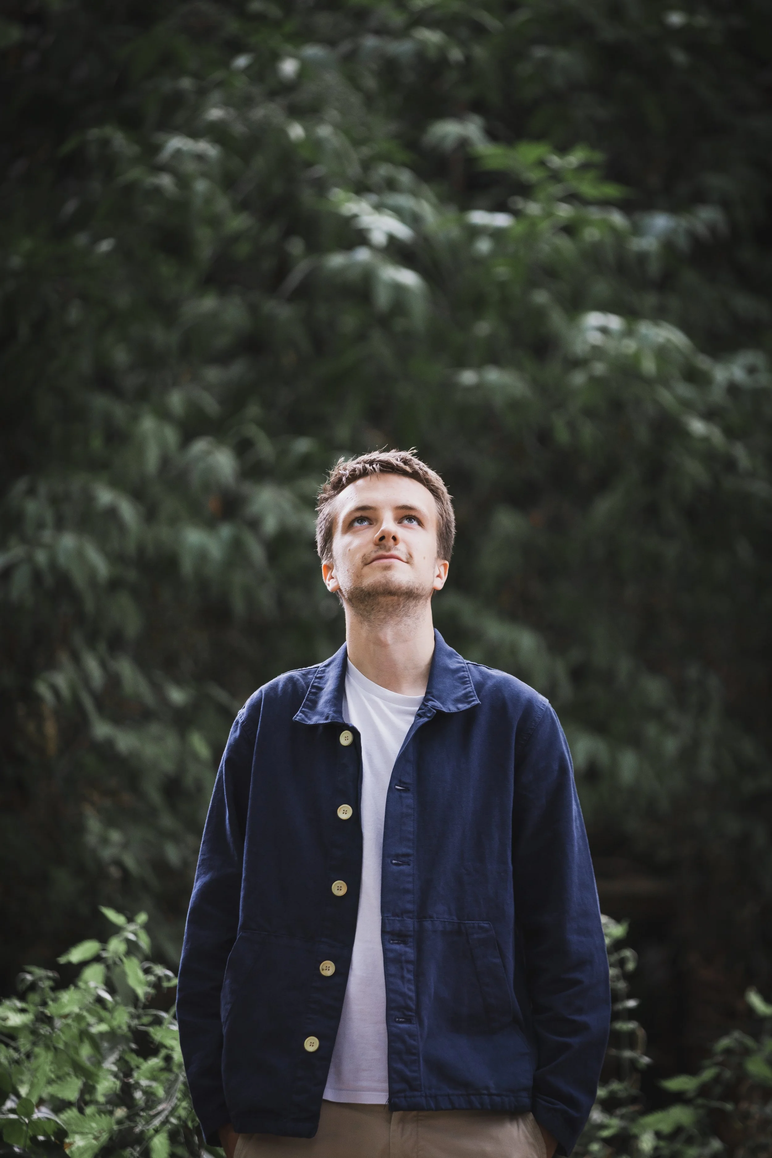 A young man stands outdoors in front of dense green foliage, looking slightly upward with a neutral expression. He is wearing a dark blue jacket over a white T-shirt and light beige pants.
