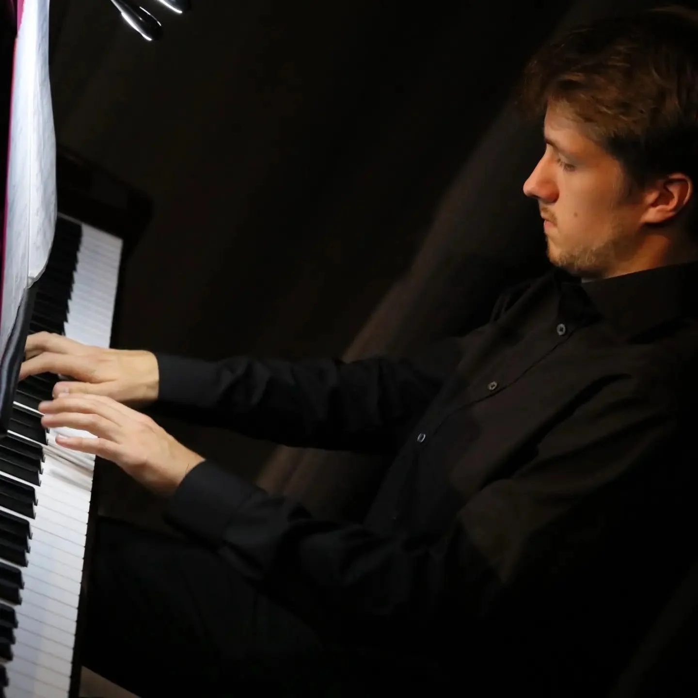 A young man playing a black grand piano, dressed in a black shirt, focusing on the piano keys.