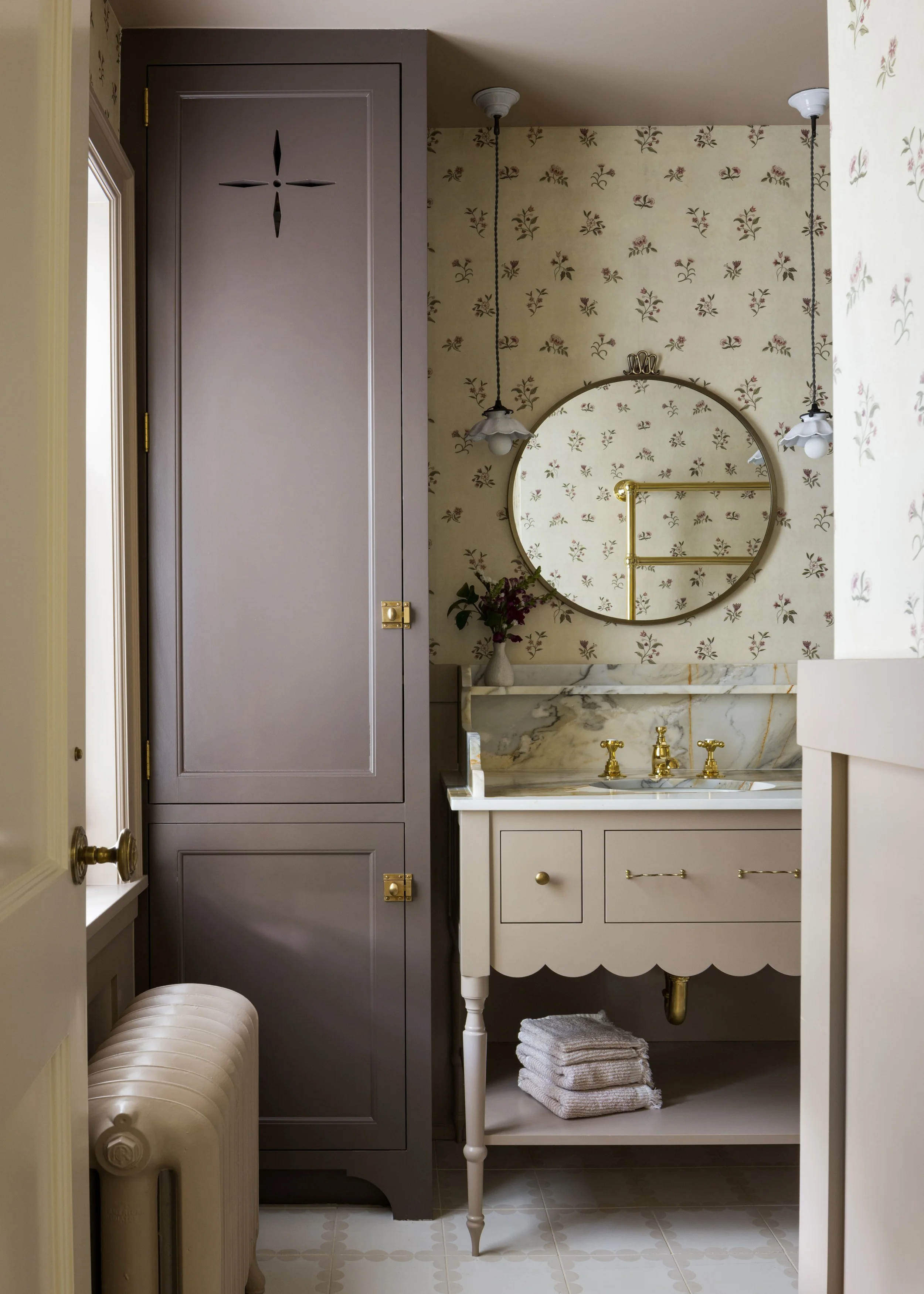 A vintage-style bathroom with a round mirror, a white vanity with gold fixtures, a vase with flowers, a tall gray cabinet, and hanging pendant lights.
