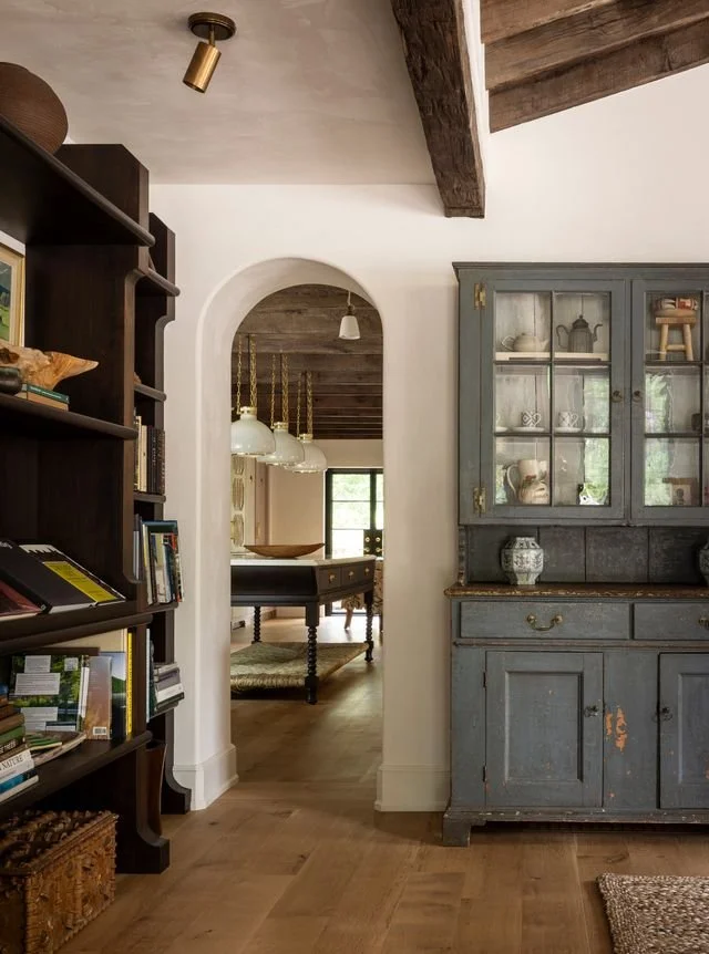 Interior view of a cozy home with wooden floors and vintage furniture, including a blue distressed sideboard and dark wood bookshelf, with an arched doorway leading to a dining area with a dark table and white pendant lights.