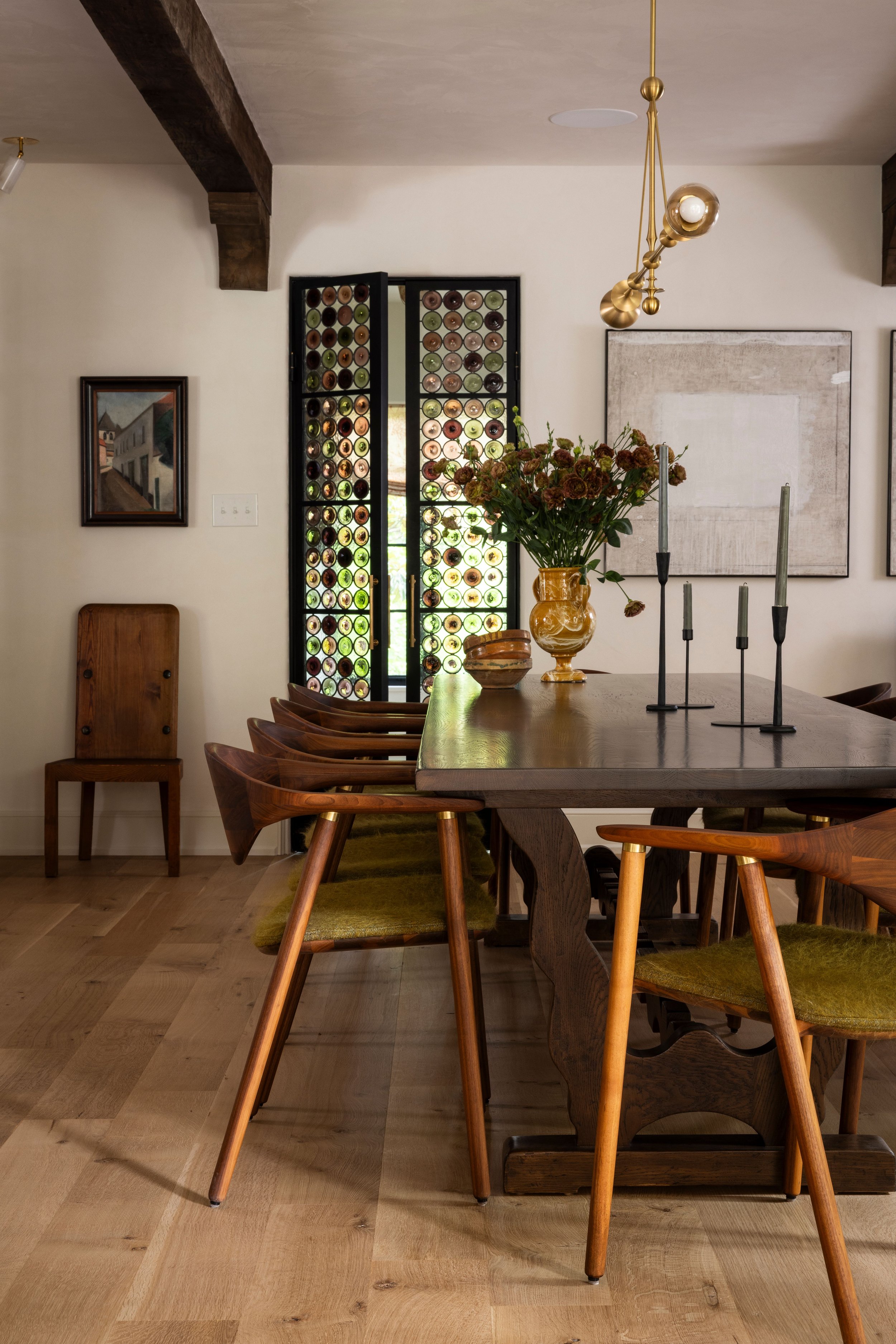 A dining room with a wooden table and chairs, a bouquet of flowers in a yellow vase, candlesticks, a large abstract painting on the wall, a window with a decorative wine bottle grid, and a modern chandelier.