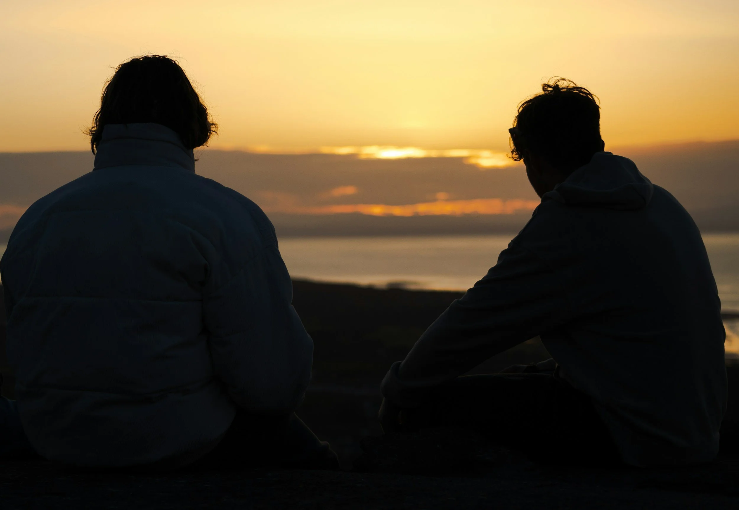 Two people sitting side by side on a beach during sunset.