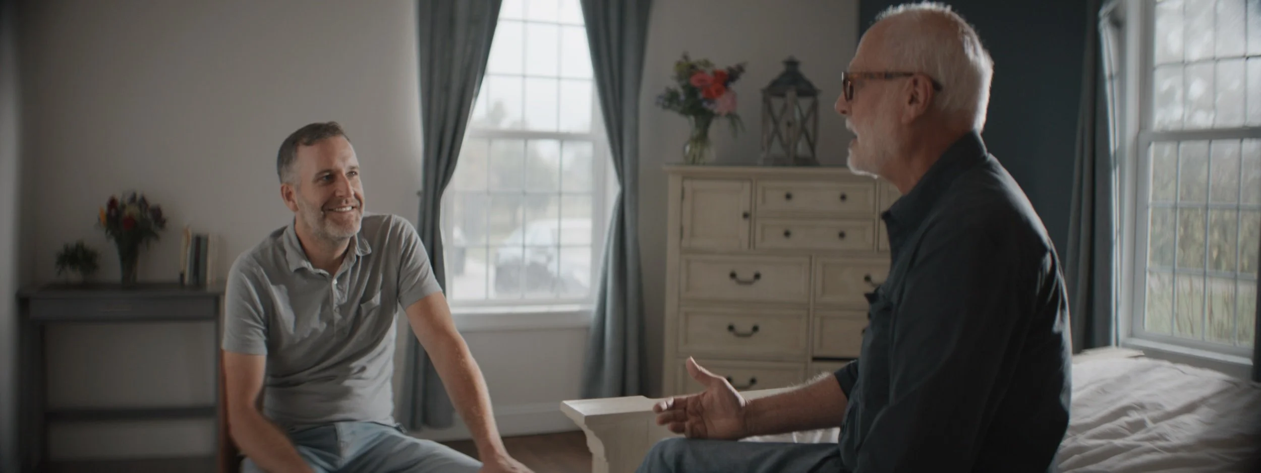 Two men sitting and talking in a bedroom with natural light, one younger and one older, both smiling.