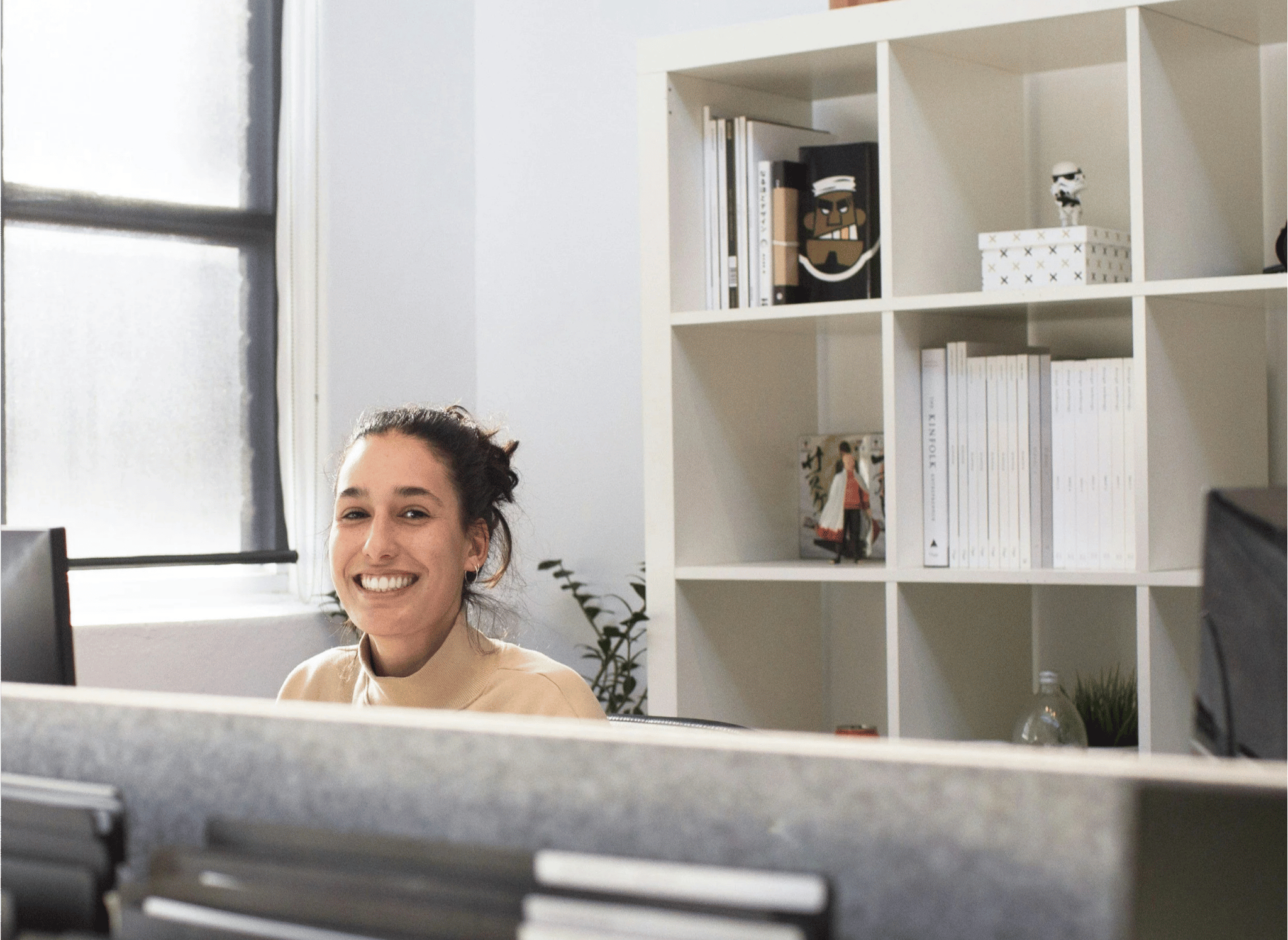 A woman receptionist smiling at her desk in a mental health office with white bookshelves in the background.