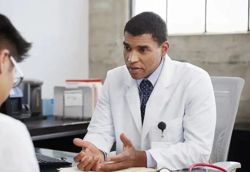 A male doctor wearing a white coat and tie speaking to a patient in an office setting.
