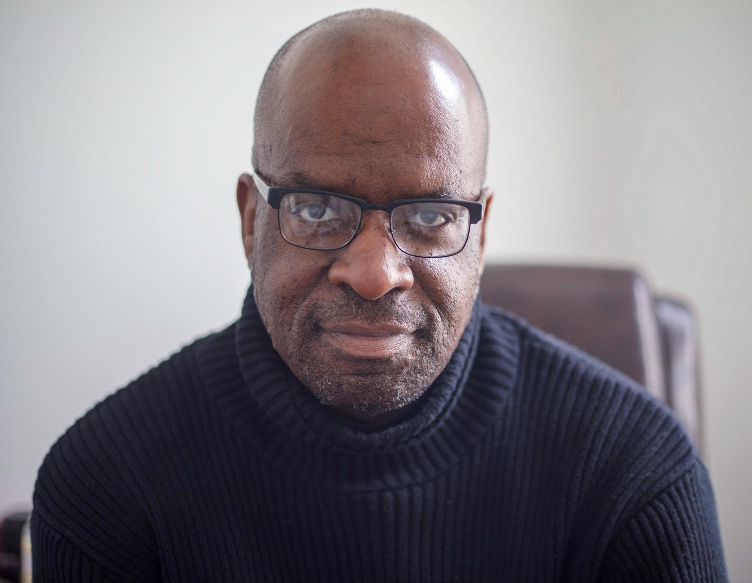 A close-up portrait of a man with a bald head, wearing glasses and a dark ribbed sweater.