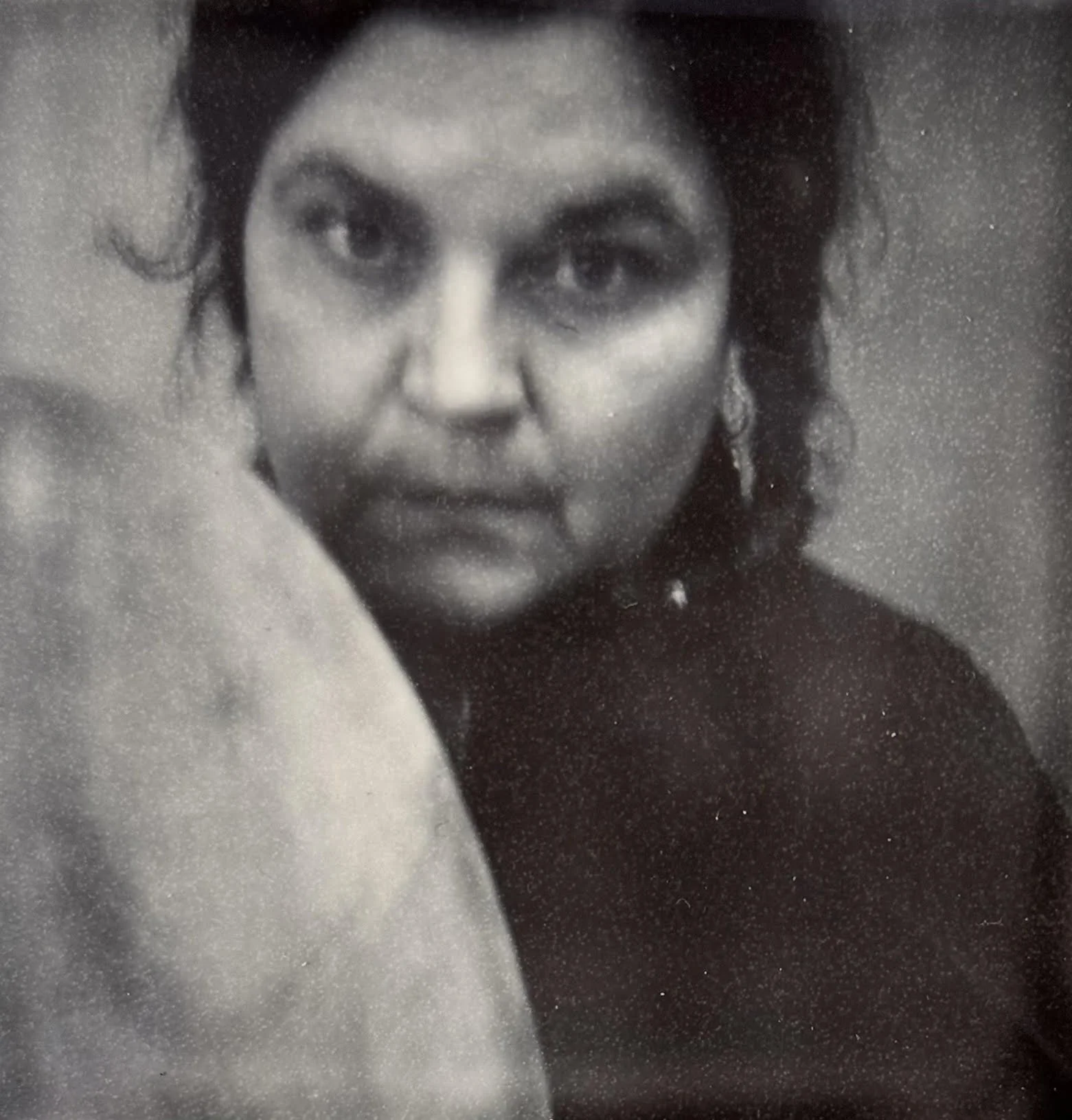 Close-up black-and-white photograph of a woman with dark hair styled with volume, wearing earrings, and a dark top with a high neckline, looking at the camera.