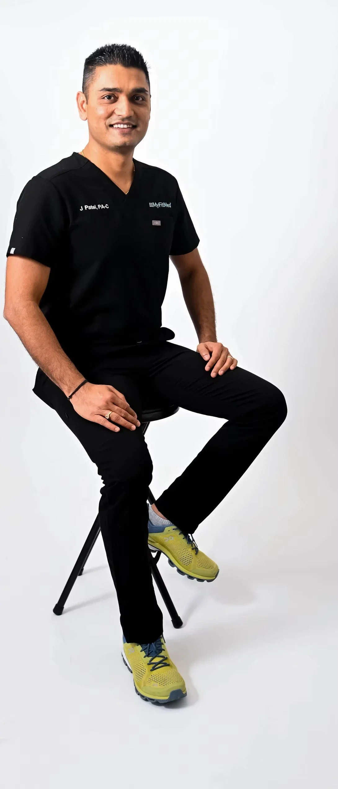 Smiling male healthcare professional in black scrubs sits on a stool.
