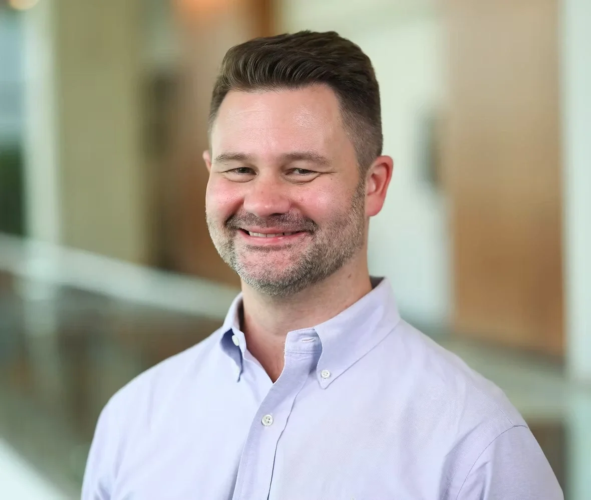 Smiling bearded man with short dark hair in a lavender button-down shirt.